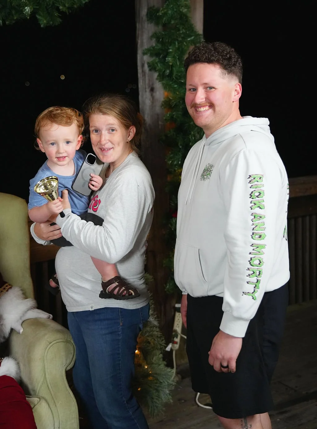 A woman holding a young boy with red hair and blue eyes, who is holding a small trophy, standing next to a man wearing a white hoodie. The woman and the boy are smiling, and the scene is decorated with greenery and lights, suggesting a holiday settin