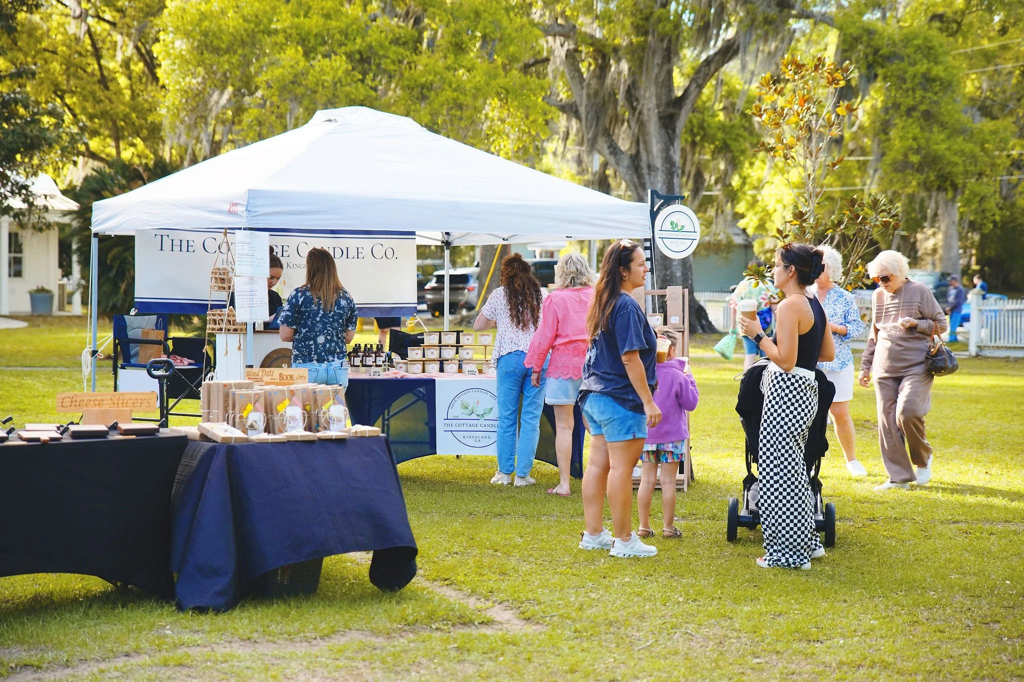 People browsing and shopping at an outdoor market under a white canopy tent with trees and grass in the background.