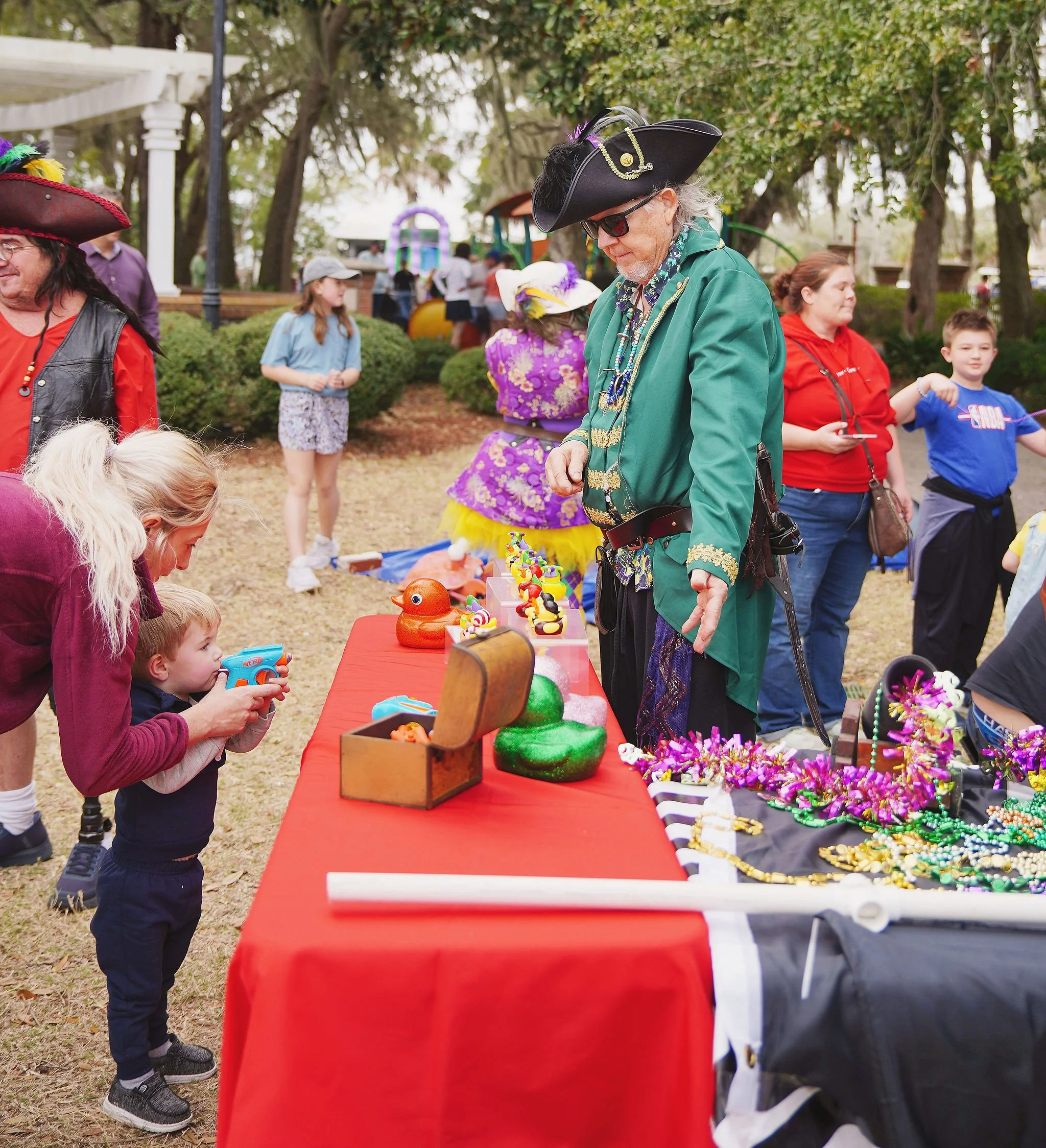 People dressed in pirate costumes at a festive outdoor event, browsing a table with colorful toys and decorations; children and adults are visible in the background.