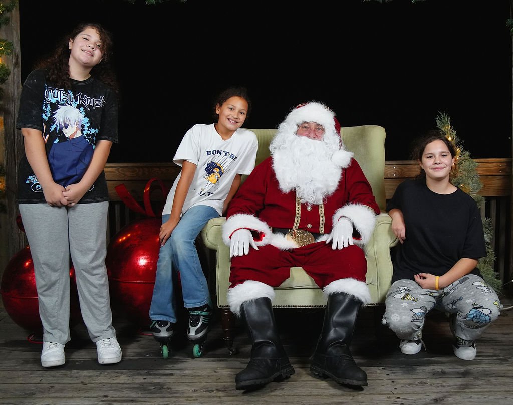 Three girls and Santa Claus sitting and standing around him. Two girls are sitting on Santa's lap, and the third girl is standing beside him. There are Christmas decorations and large red ornaments in the background.