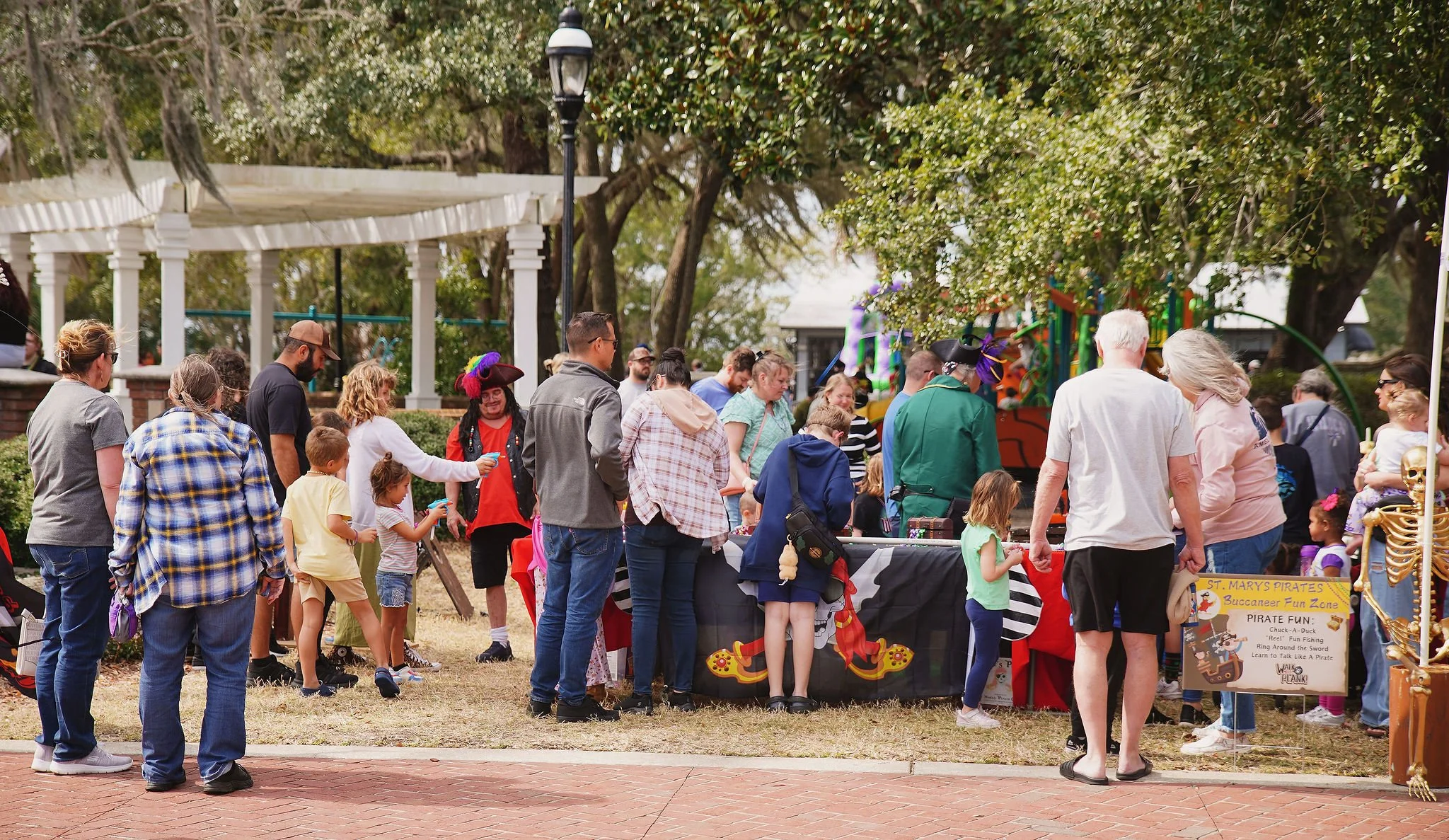 People gather around a pirate-themed game booth at an outdoor event, with children and adults standing in line. The scene is set in a park with trees, a gazebo, and decorative elements like skeletons. A sign on the booth reads 'St. Mary’s Pirates Buc