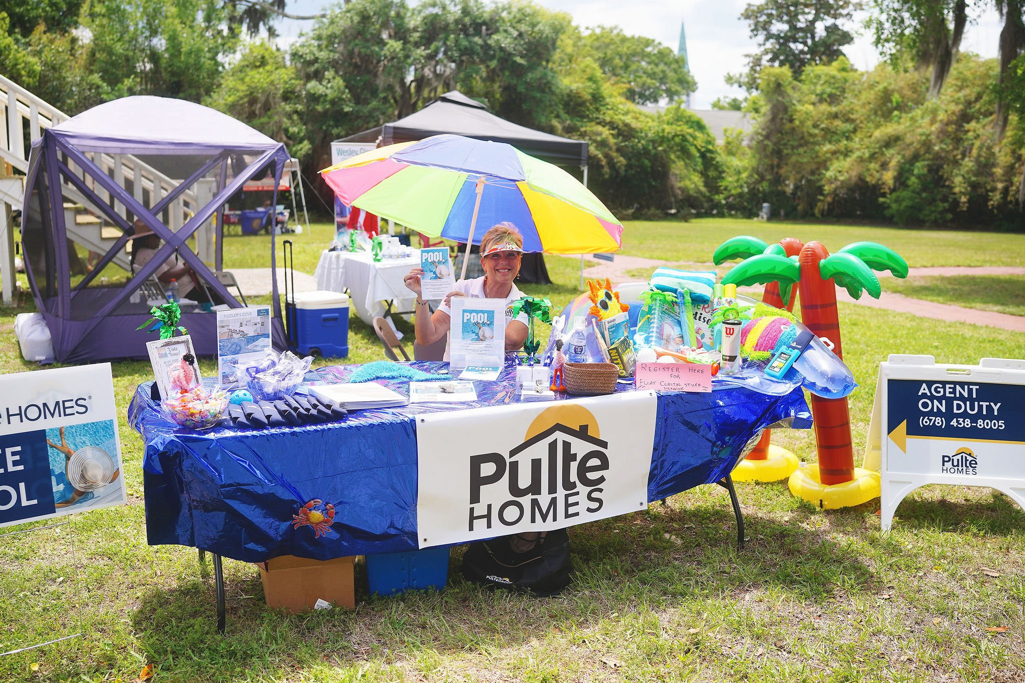 A woman sitting at a table with a Pulte Homes banner, selling pool-themed items at an outdoor event, with inflatable palm trees and a colorful umbrella, in a grassy park with trees in the background.