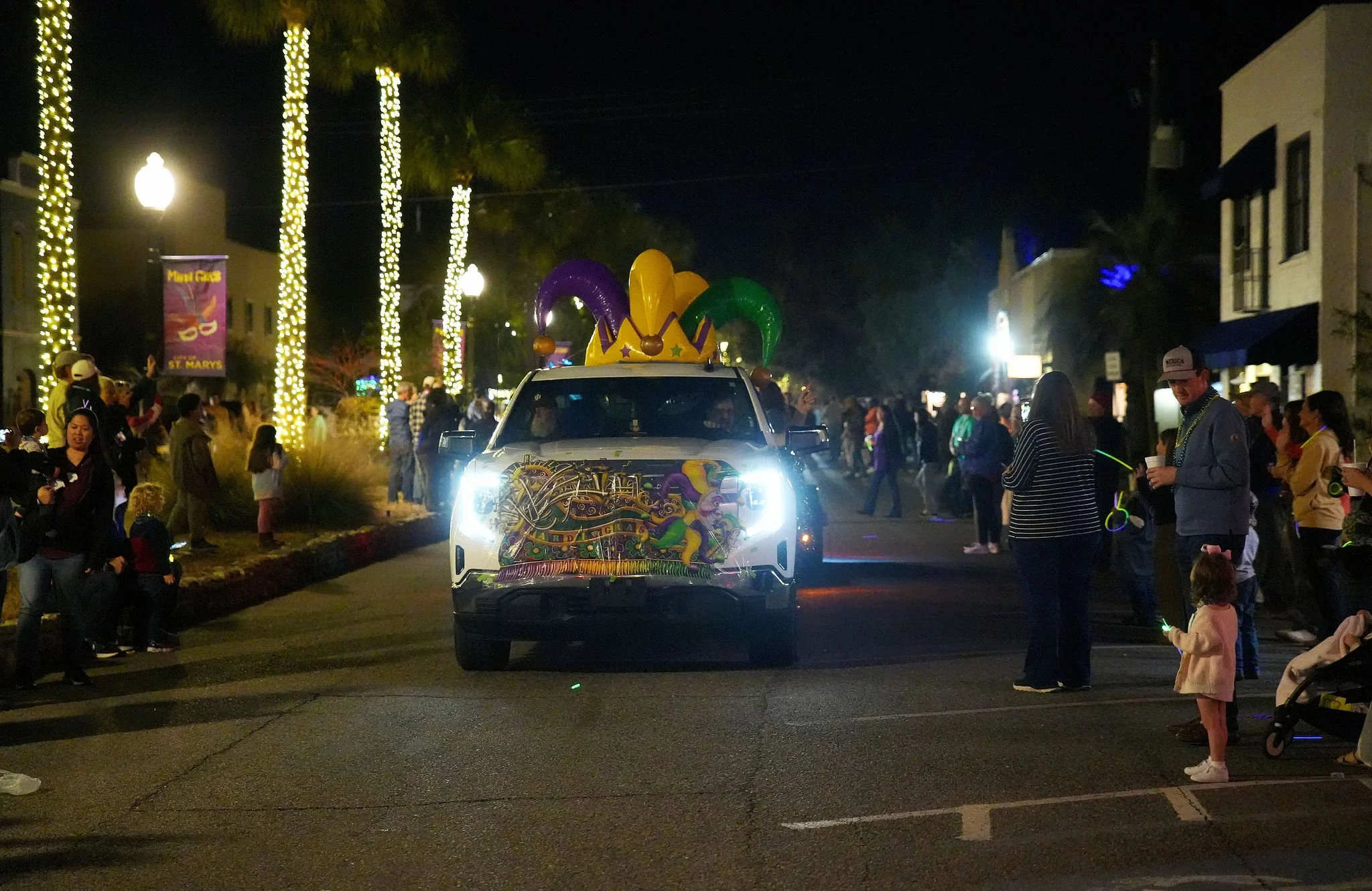 A parade float with colorful jester hats in the middle of a city street at night, surrounded by people watching the parade, with trees decorated with string lights.
