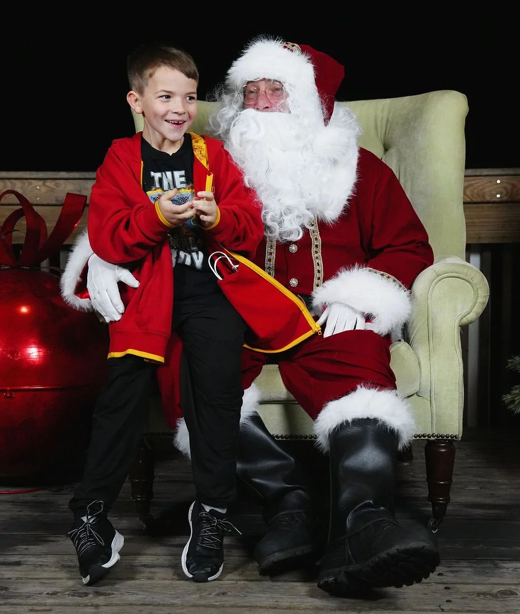 A young boy sitting on Santa Claus's lap, smiling, holding a small gift and wearing a red jacket, with Santa dressed in his traditional red suit, white beard, and hat, sitting on a yellow armchair.