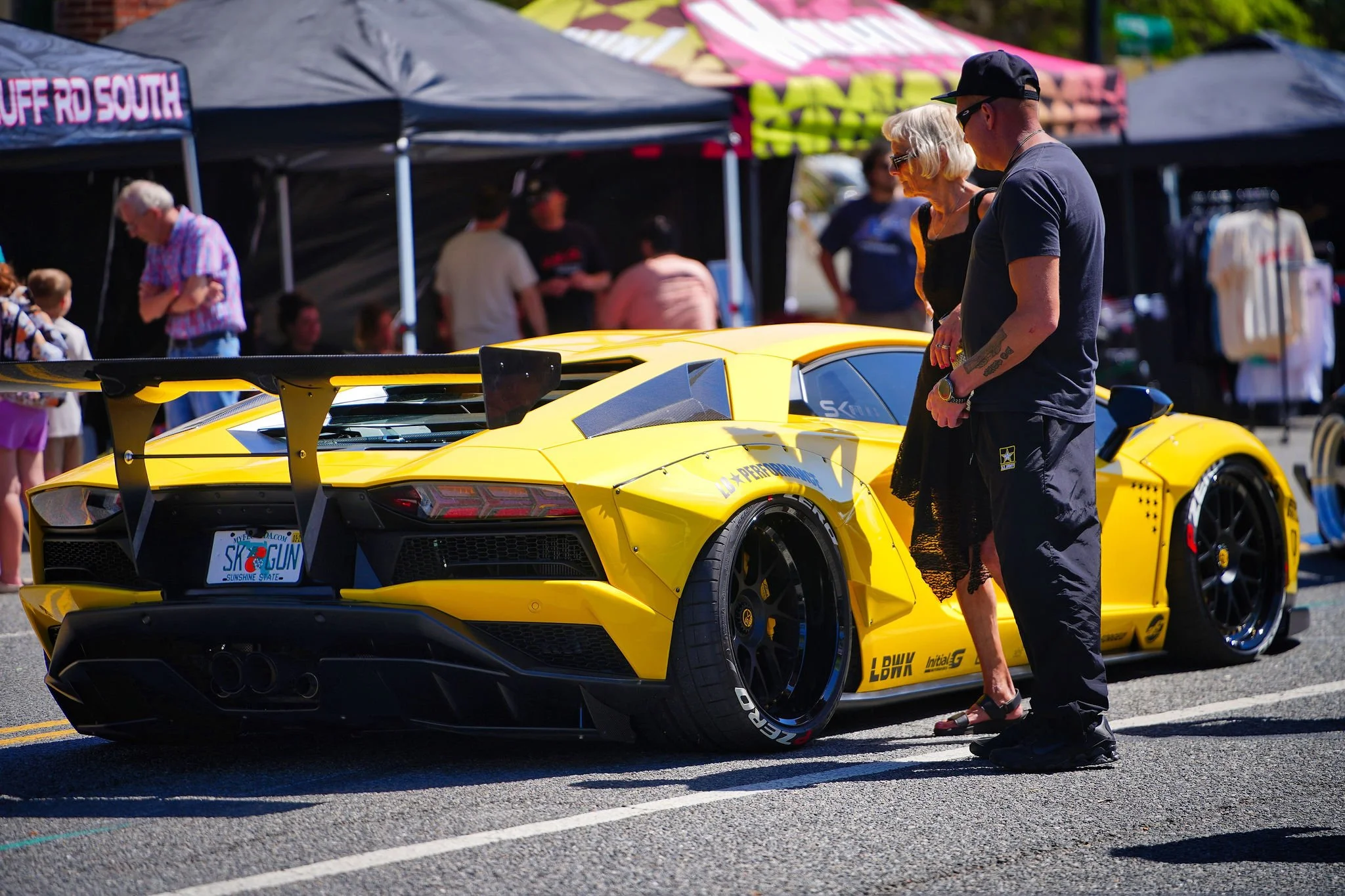 A bright yellow Lamborghini sports car with a large rear spoiler at an outdoor car show. Two people, a woman with blonde hair and sunglasses and a man with tattoos wearing a black shirt and cap, are looking at the car. There are tents and other peopl
