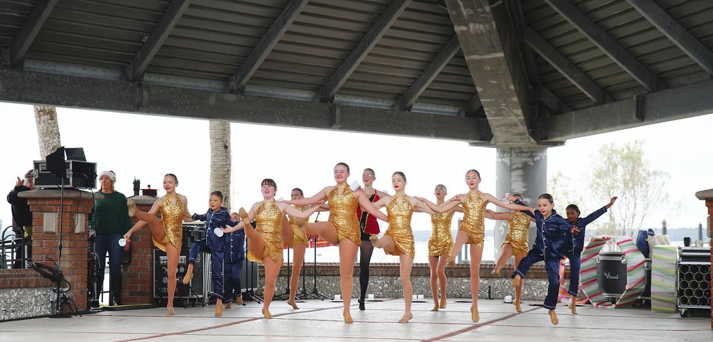 Group of young girls performing a dance on a stage under a pavilion, wearing matching golden costumes and black or navy blue tracksuits.