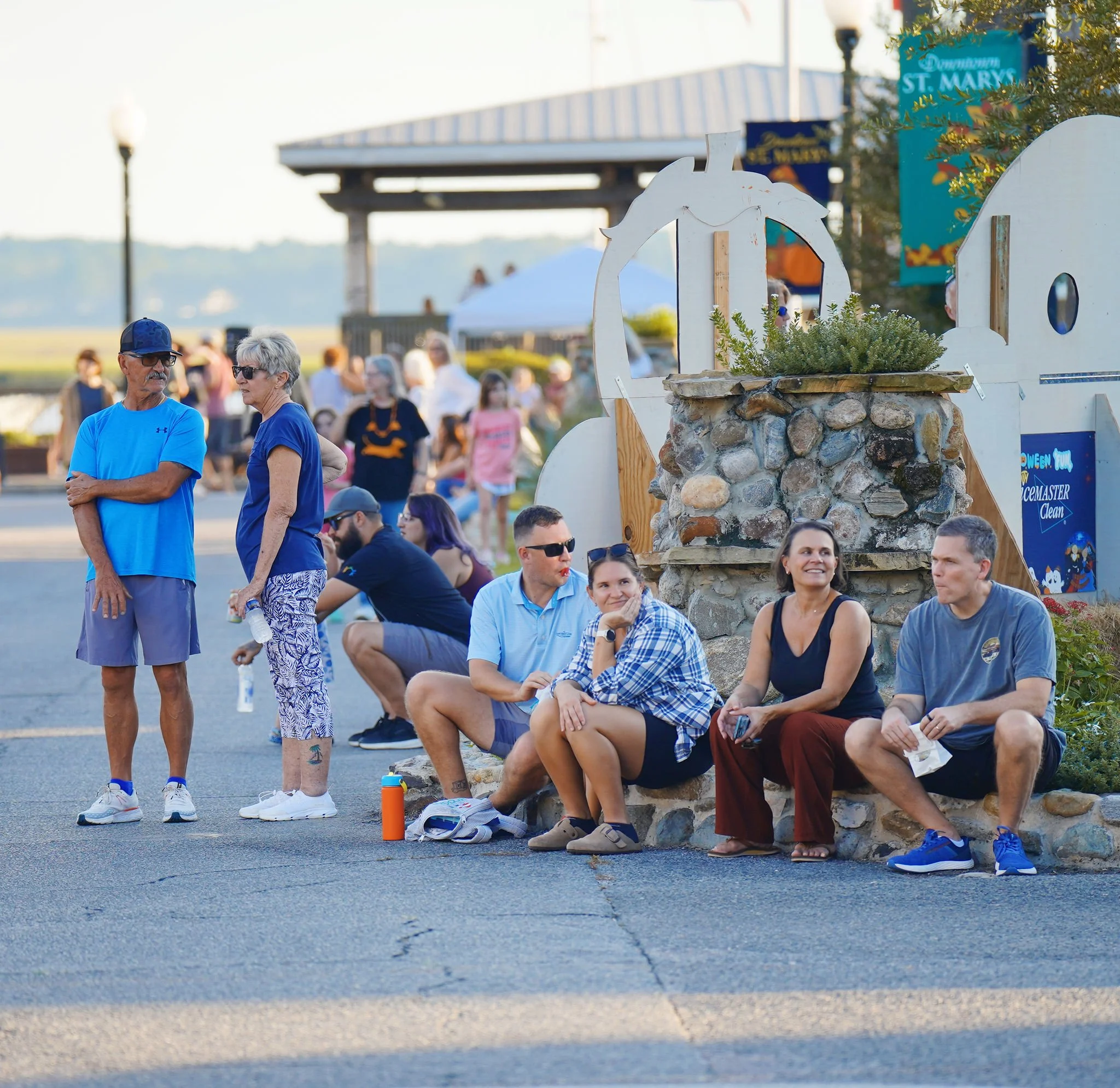 People gathered outdoors, some sitting on the ground and others standing, near a decorative stone structure with a white wood cutout of a horse's head.
