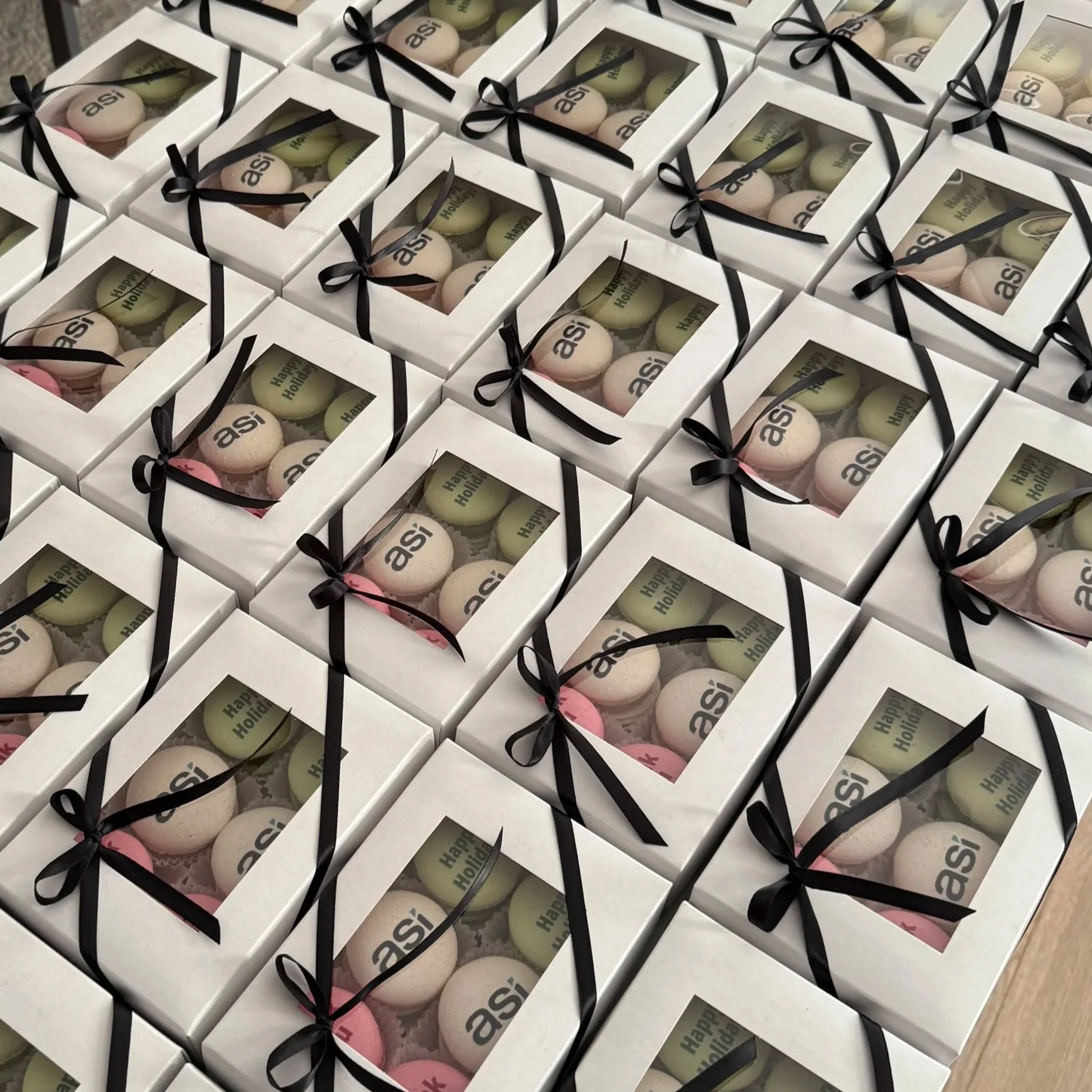 Multiple white gift boxes with transparent windows, each containing pastel-colored macarons with words like "asi" and "Happy Holidays" printed on them. The boxes are decorated with black ribbons.