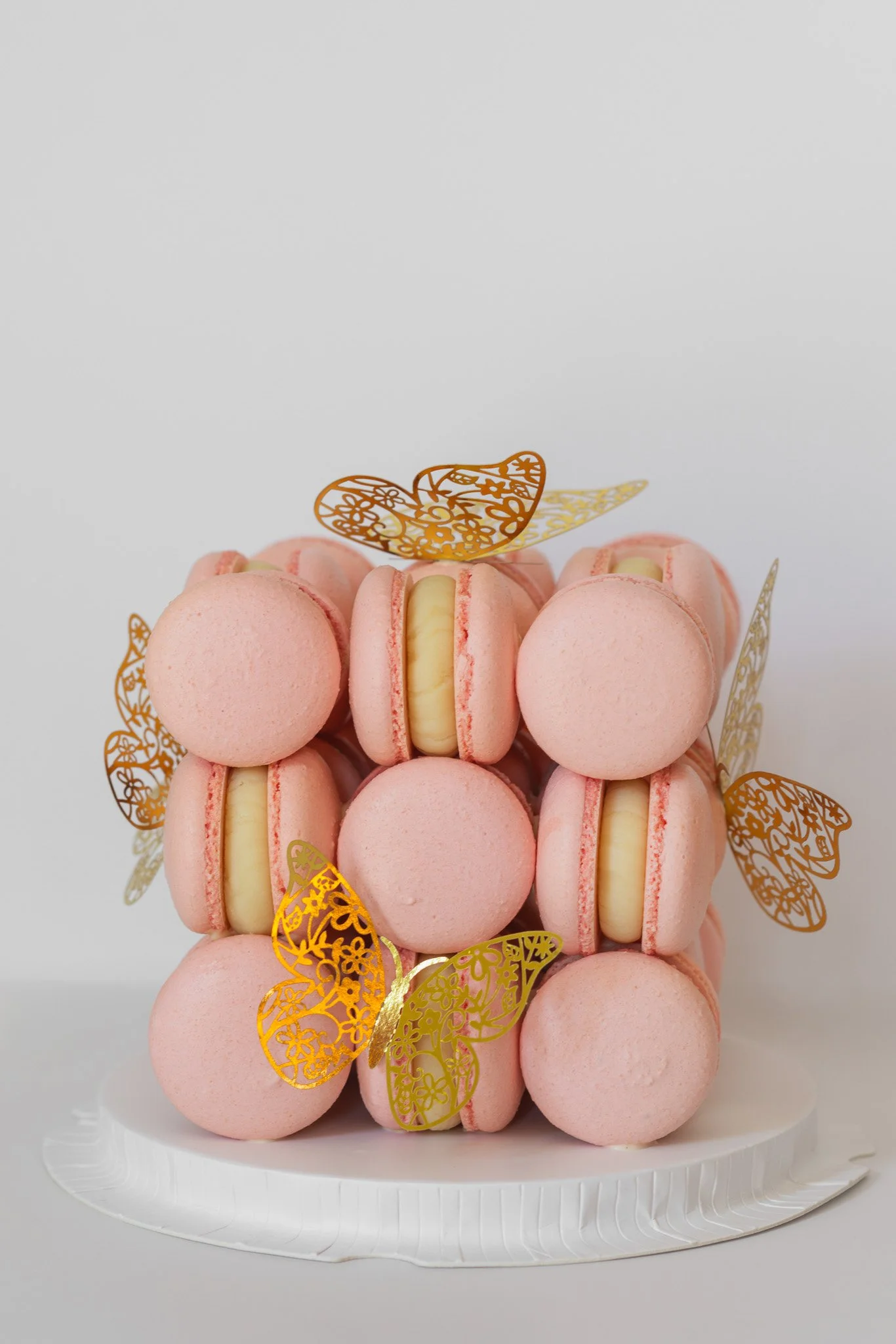 A cake made of pink macarons with a white filling, decorated with gold butterfly toppers, on a white paper plate.