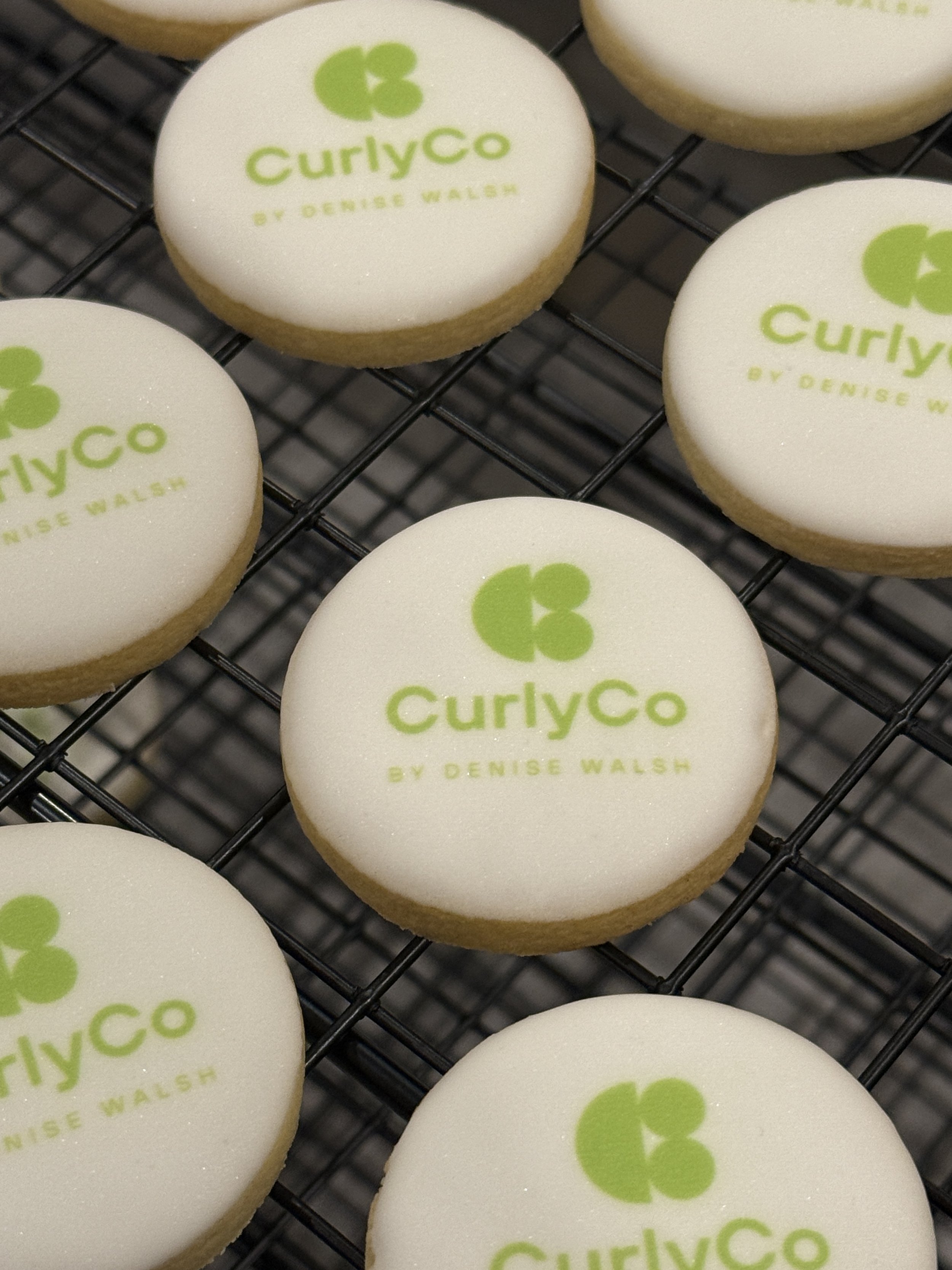 Cookies with green logo and text that reads 'CurlyCo by Denise Walsh' on white icing, resting on a black wire cooling rack.
