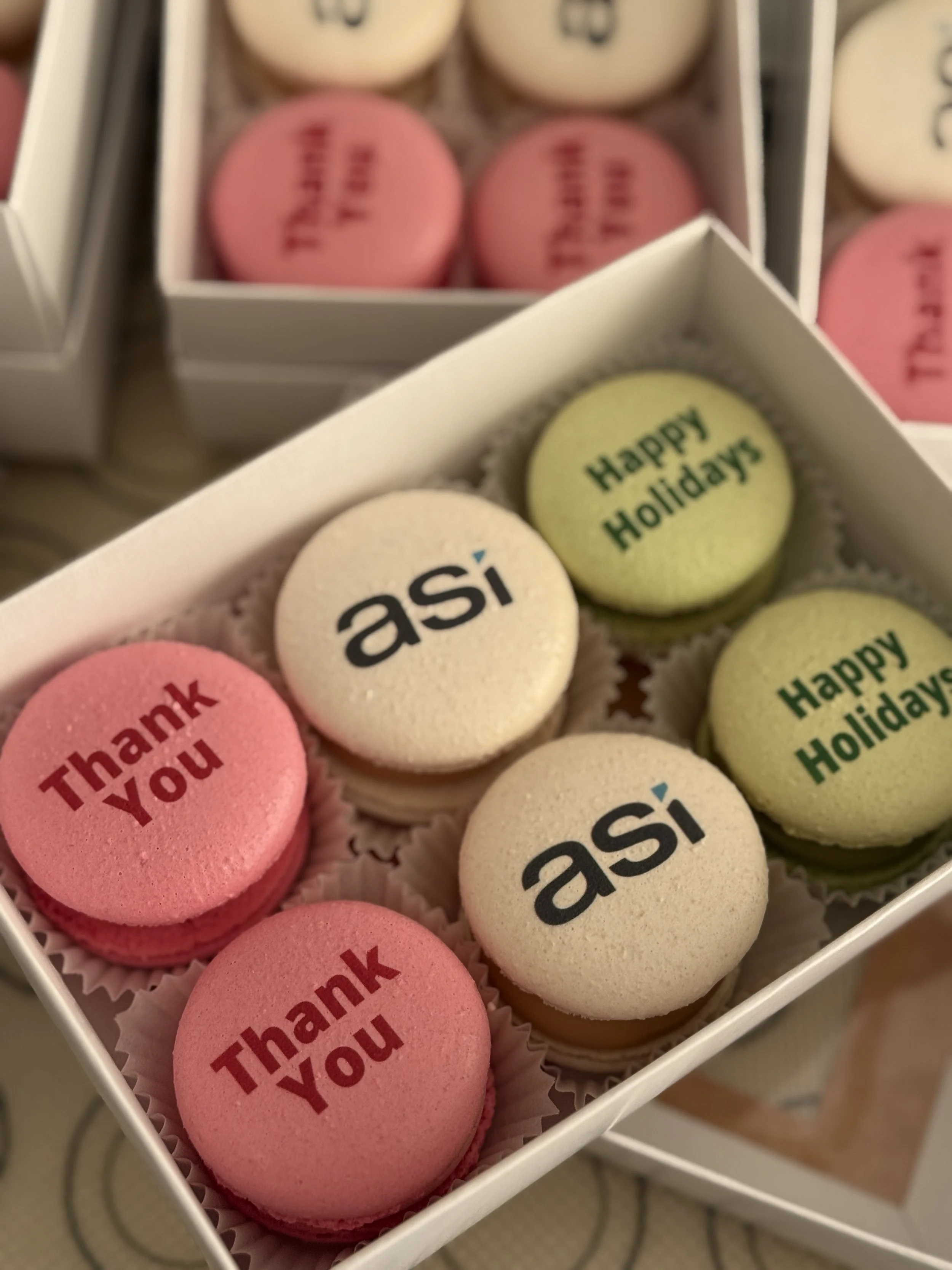 Assorted macarons in a box with messages including "Thank You," "Happy Holidays," and "asi."