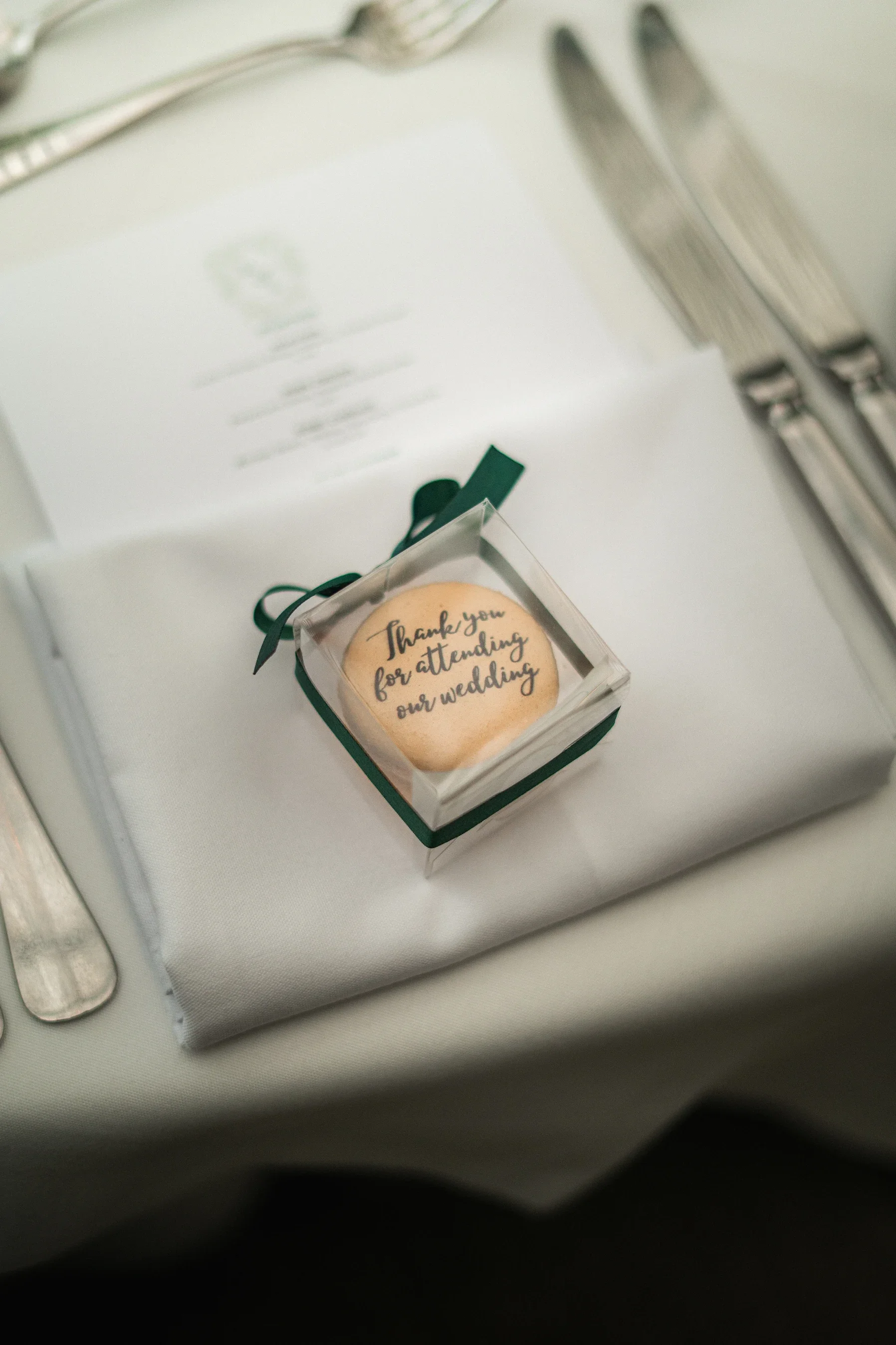 A wedding favor featuring a cookie with the message 'Thank you for attending our wedding' in a small transparent box with a green ribbon, placed on a white tablecloth beside silverware and a menu.