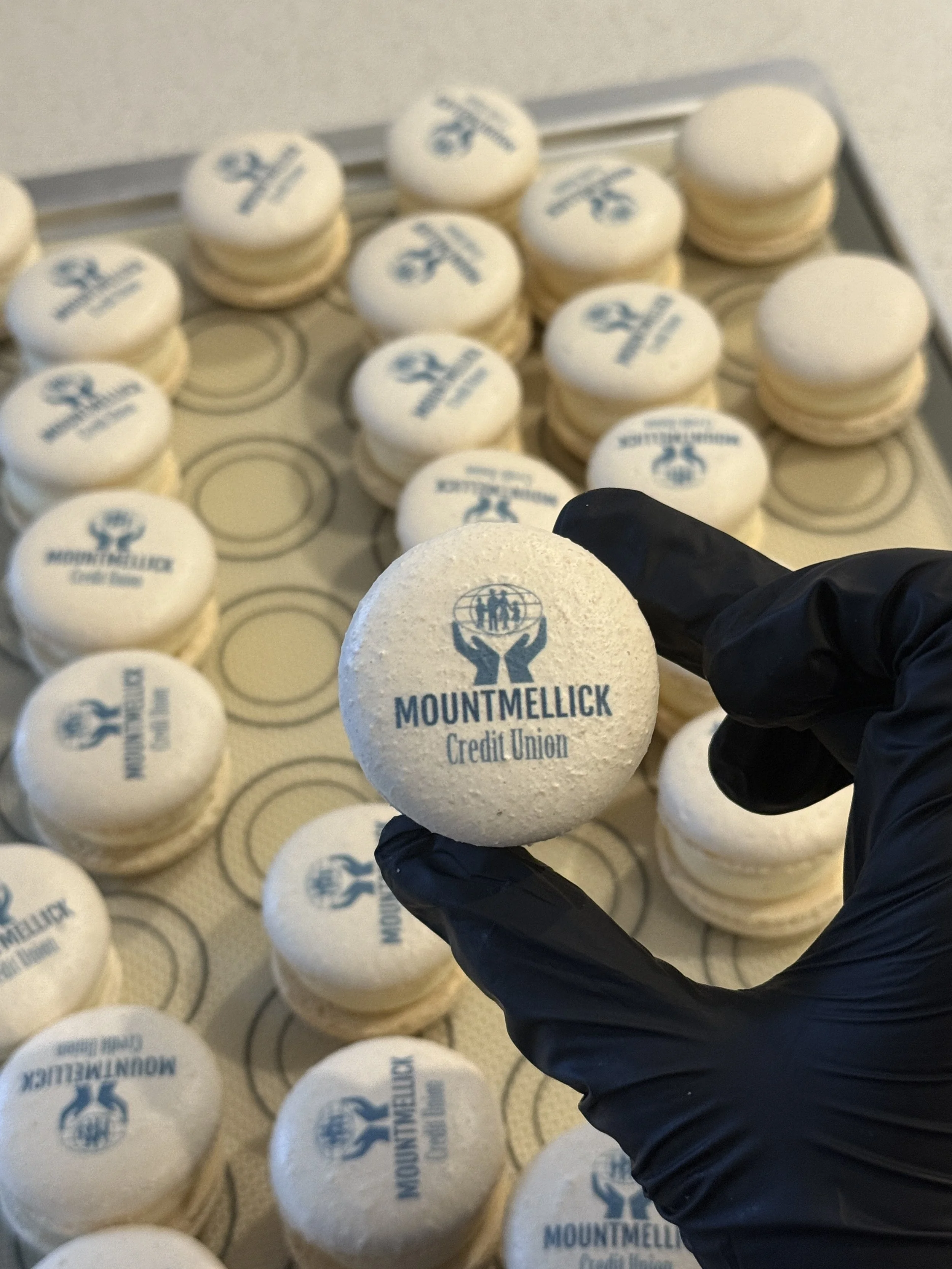A person wearing black gloves holding a white foam ball with 'Mountmellick Credit Union' printed on it in blue. Several similar foam balls are arranged on a baking tray in the background.