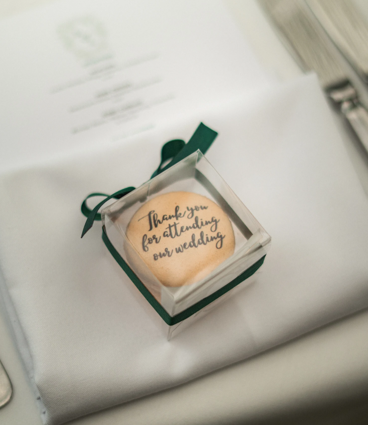 cream macaron with black printed text on it saying 'thank you for attending our wedding' in a clear box with a green ribbon