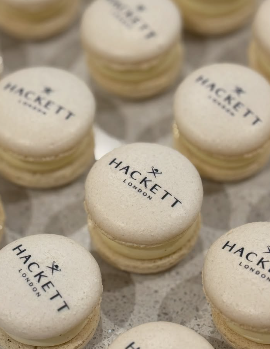 Multiple white macarons with black 'HACKETT LONDON' logos on top, arranged on a surface.
