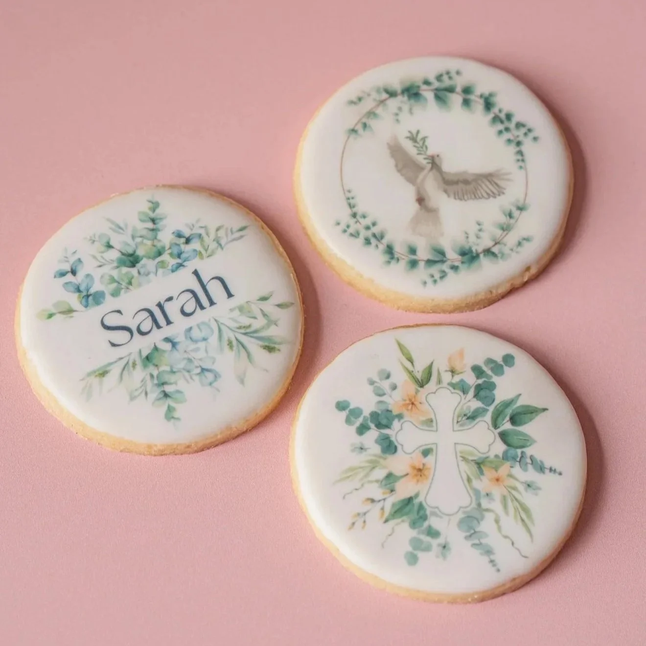 Three decorated cookies on a pink surface, one with the name 'Sarah' and floral design, another with a dove within a wreath, and the third with a cross and floral design.