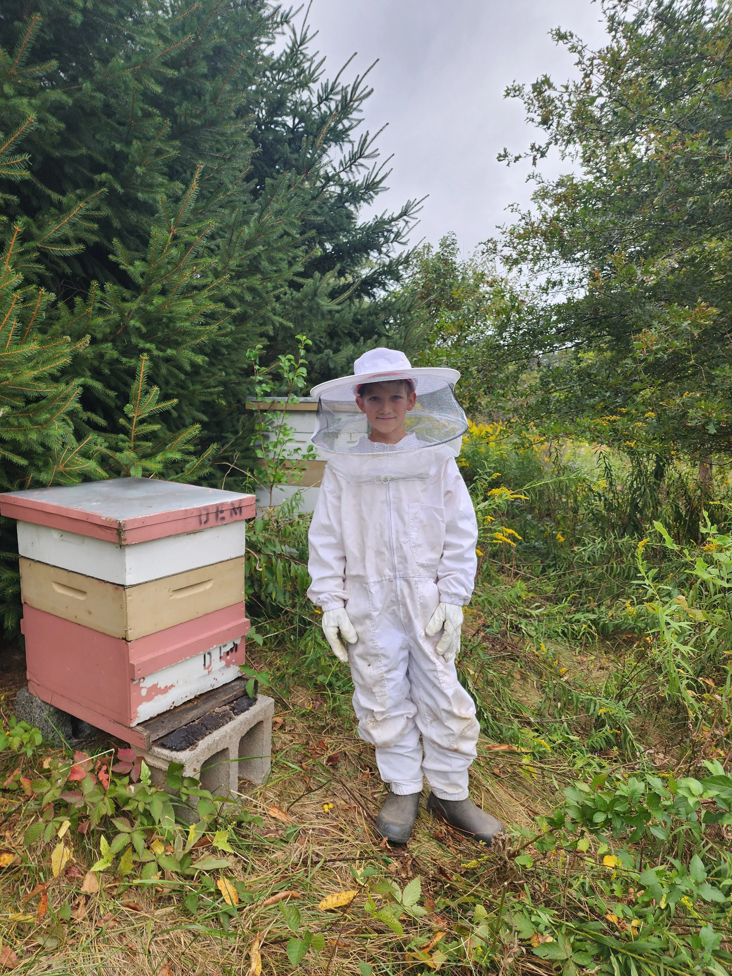 A young boy in a white bee suit standing by a bee hive.
