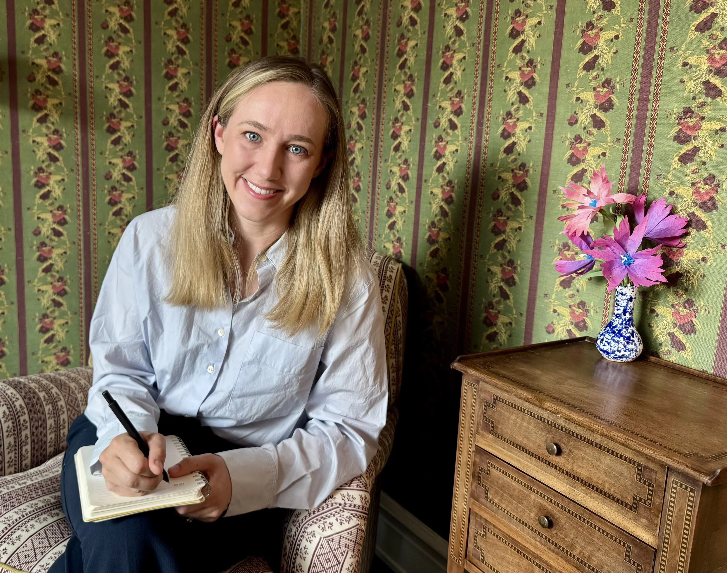 A smiling woman with blonde hair and blue eyes sits on a patterned sofa, holding a pen and notebook, in a room with green patterned wallpaper and a wooden side table with a blue and white vase containing pink flowers.