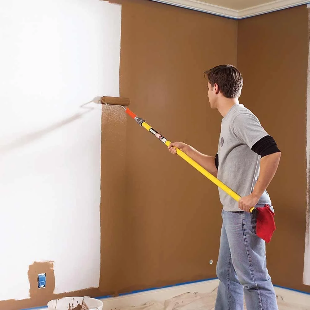 A man painting a wall with a roller, applying white paint over a brown wall in a room.