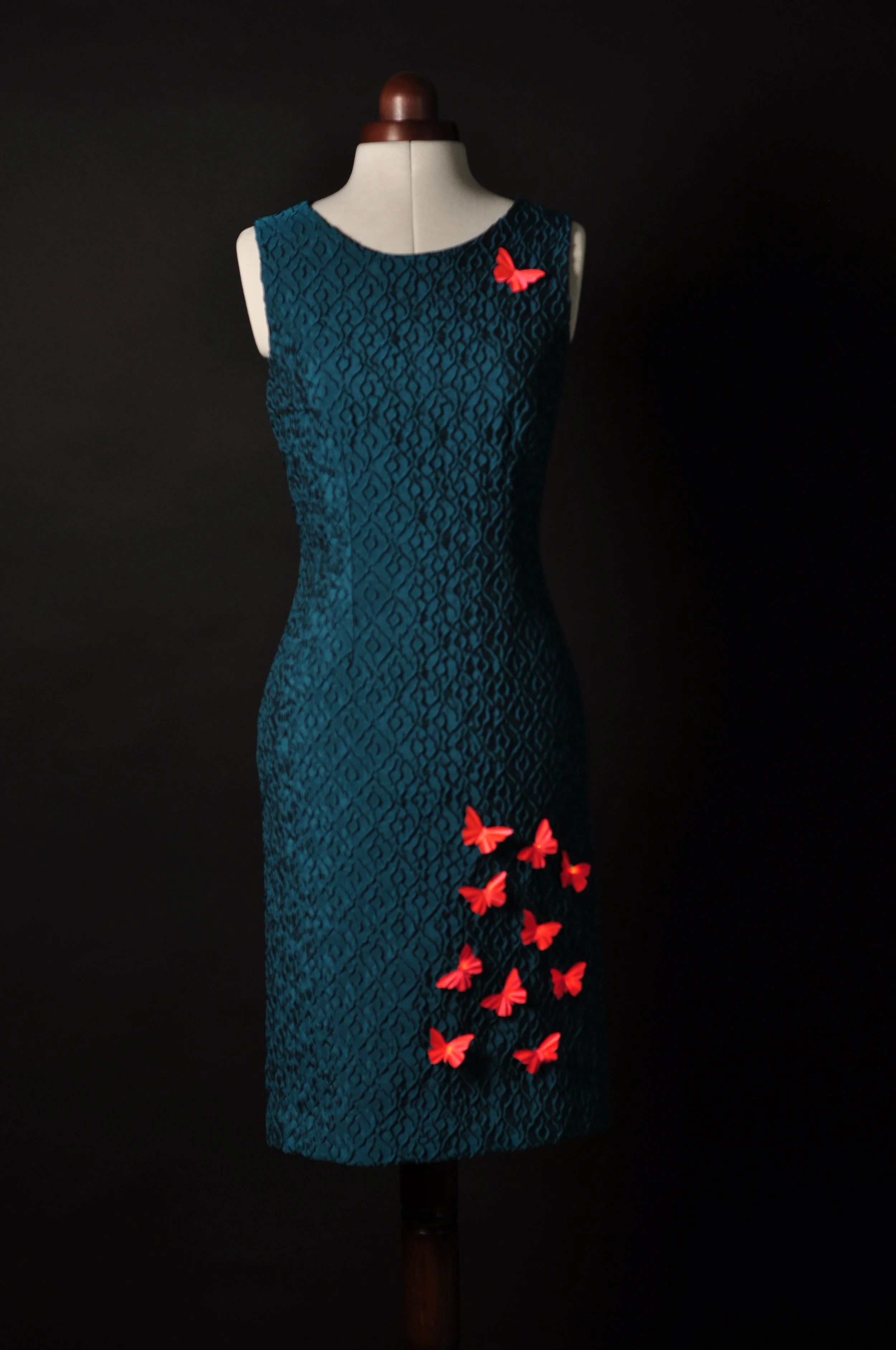 Blau-grünes Etuikleid aus Piqué-Stoff mit aufgesetzten orangen Schmetterlingen auf Schneiderpuppe, schwarzer Hintergrund.