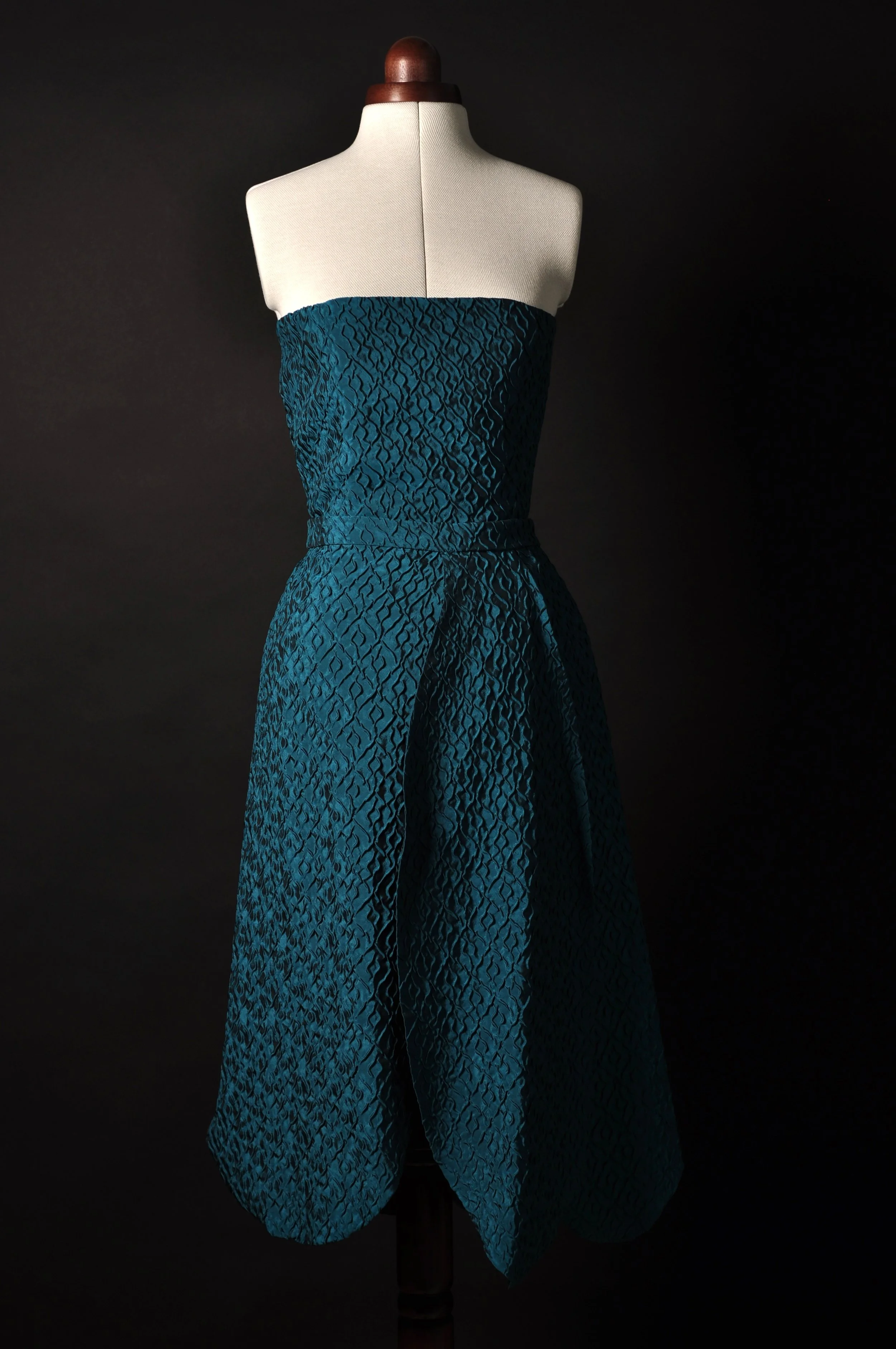 Blau-grünes, trägerfreies Kleid auf einer Schneiderpuppe vor schwarzem Hintergrund, Rock wie Tulpenblätter, Piqué-Stoff.
