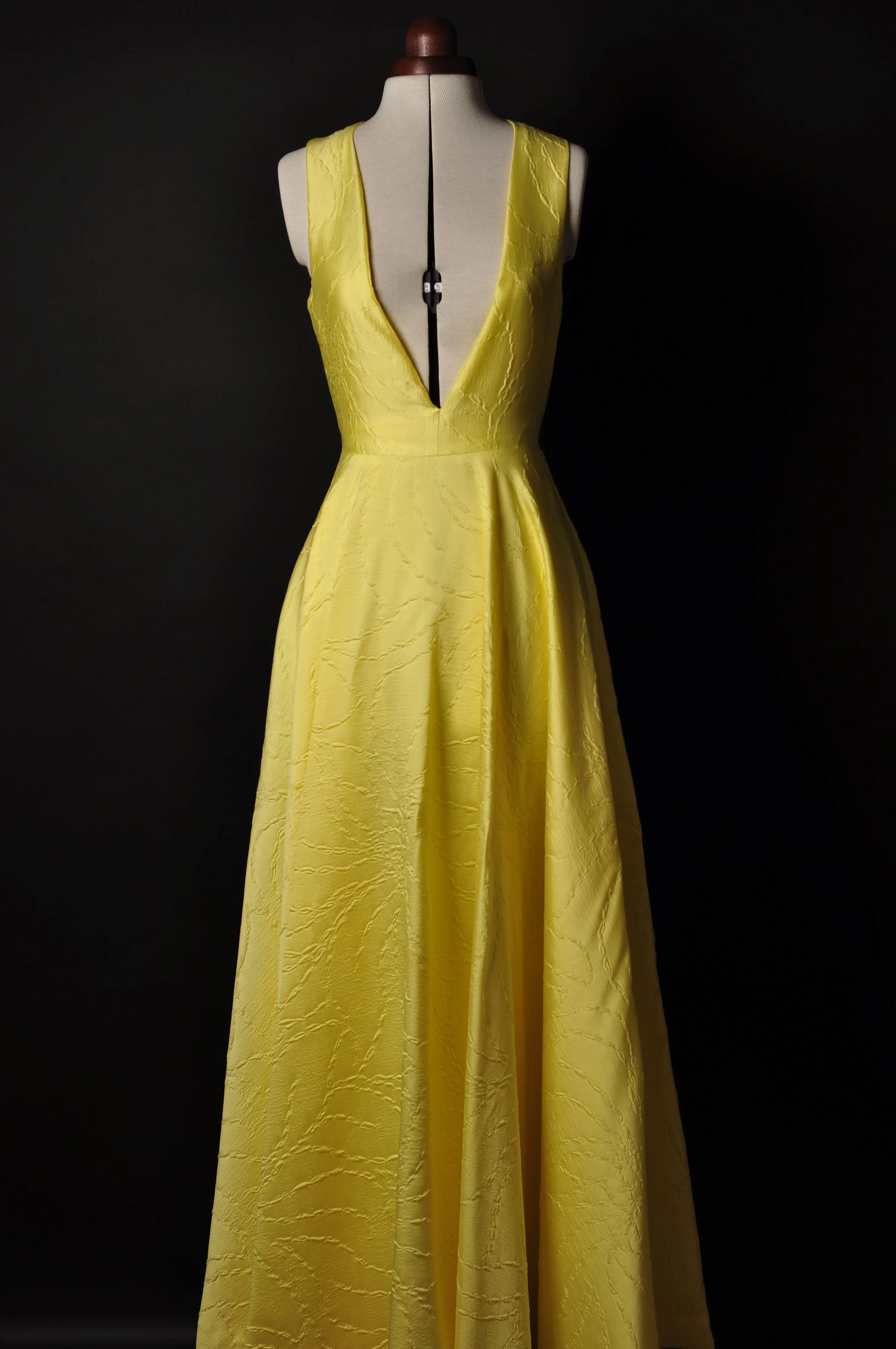 Gelbes Abendkleid aus Jacquard-Stoff mit tiefem Ausschnitt vorne auf einer Schneiderpuppe vor schwarzem Hintergrund.
