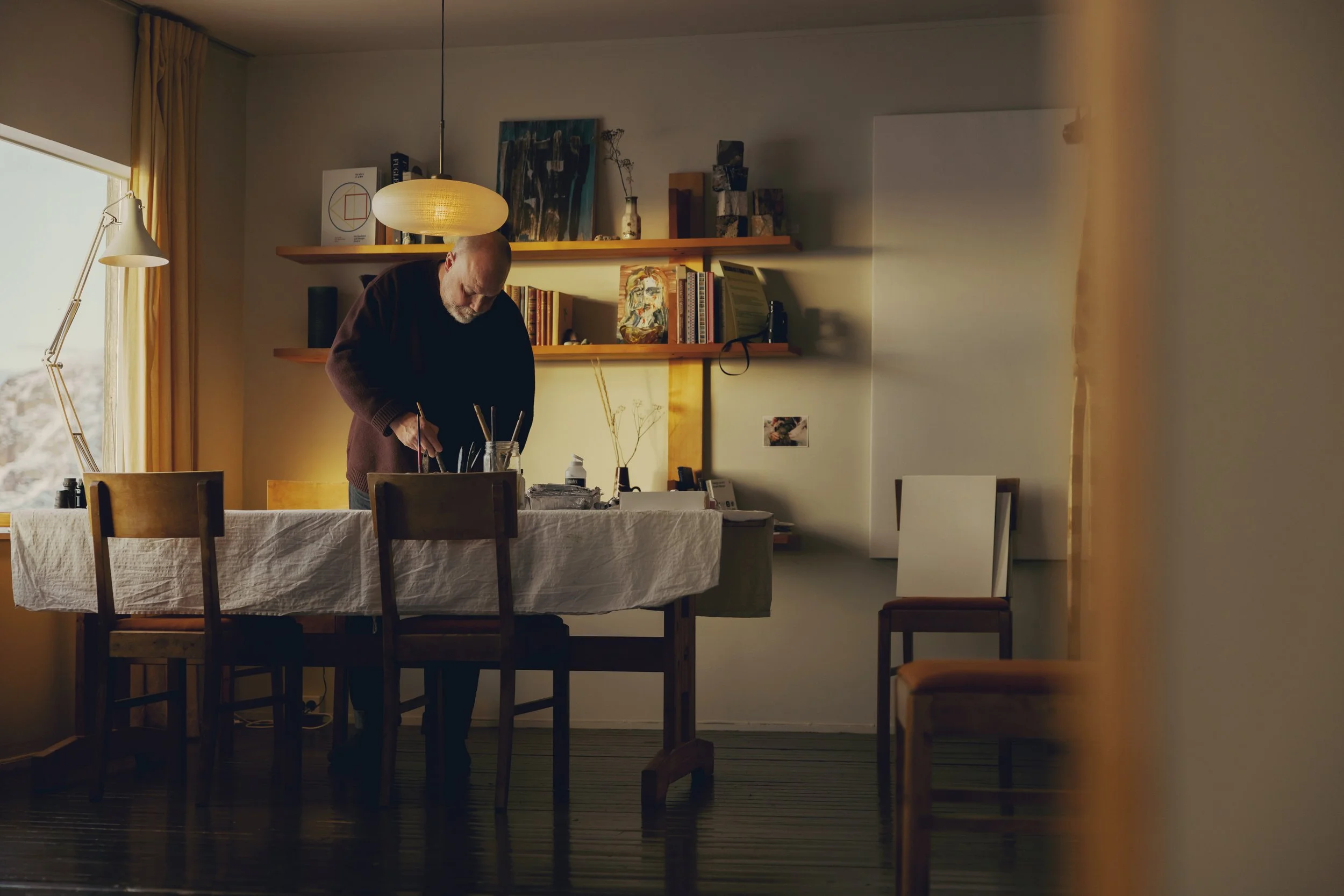A man with a beard and bald head working at a dining table covered with a white cloth, in a warmly lit room with a large window with yellow curtains. The room has artwork, books, and decorative items on shelves. There is a hanging lamp above the table and a lamp with an adjustable arm near the window.