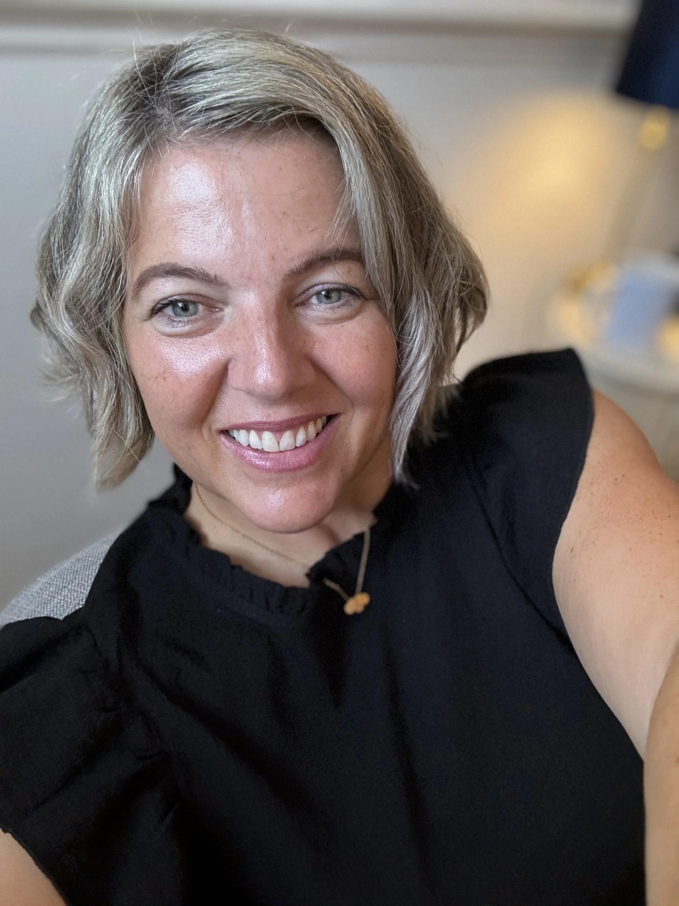 A smiling woman with short blonde hair wearing a black top, taking a selfie indoors.