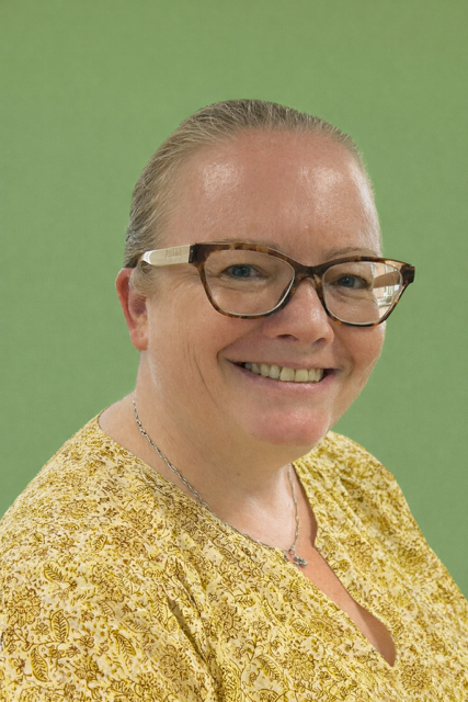 A smiling woman with glasses, wearing a beige sweater, standing indoors near green plants.
