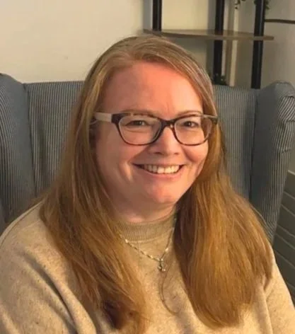 A smiling woman with glasses, wearing a beige sweater, standing indoors near green plants.