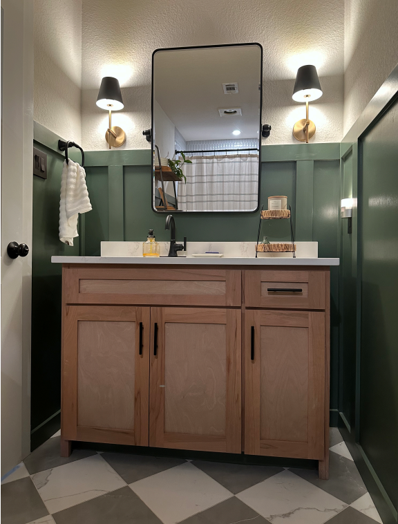 Bathroom vanity with a wooden cabinet, a rectangular mirror, two wall-mounted lamps, and a small shelf with a candle, set against green paneled walls.