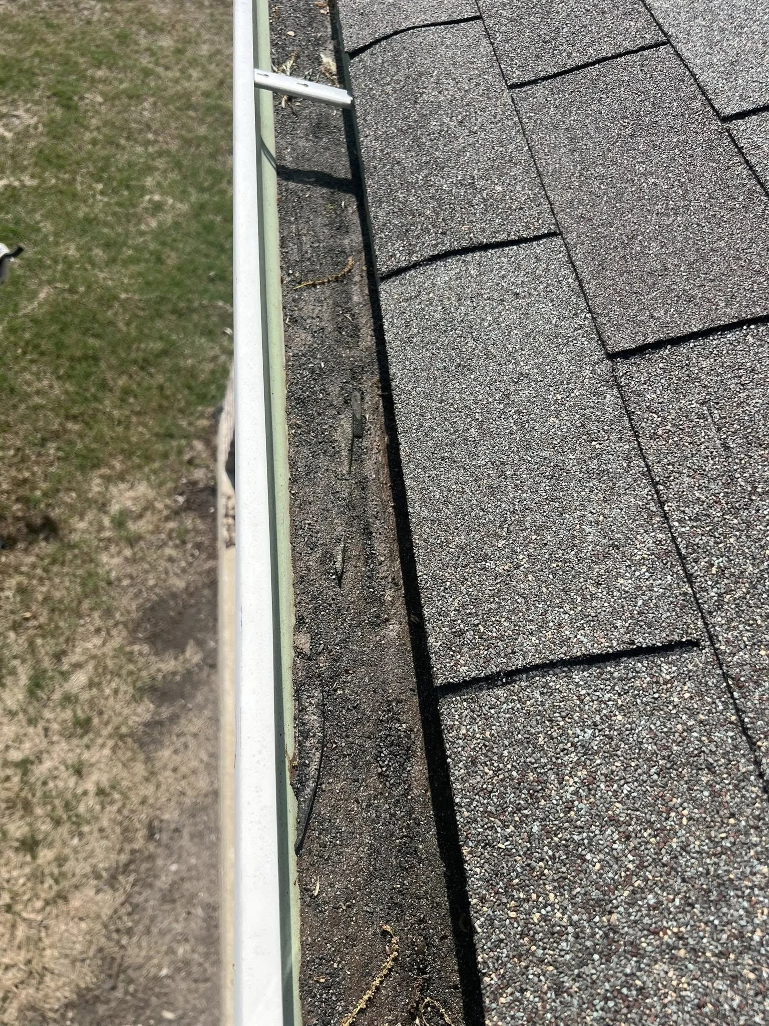 Close-up of a roof edge with gravel shingles, showing damage and separation along the seam, with a gutter and grass visible on the side.