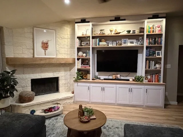 Living room with a built-in white entertainment center housing a large flat-screen TV, surrounded by shelves with books and decorative items. A stone fireplace with a wooden mantel and a picture of a tree is on the left. A small round coffee table with decorative items is in the center, with a gray area rug underneath. A potted plant is on the left near the fireplace.