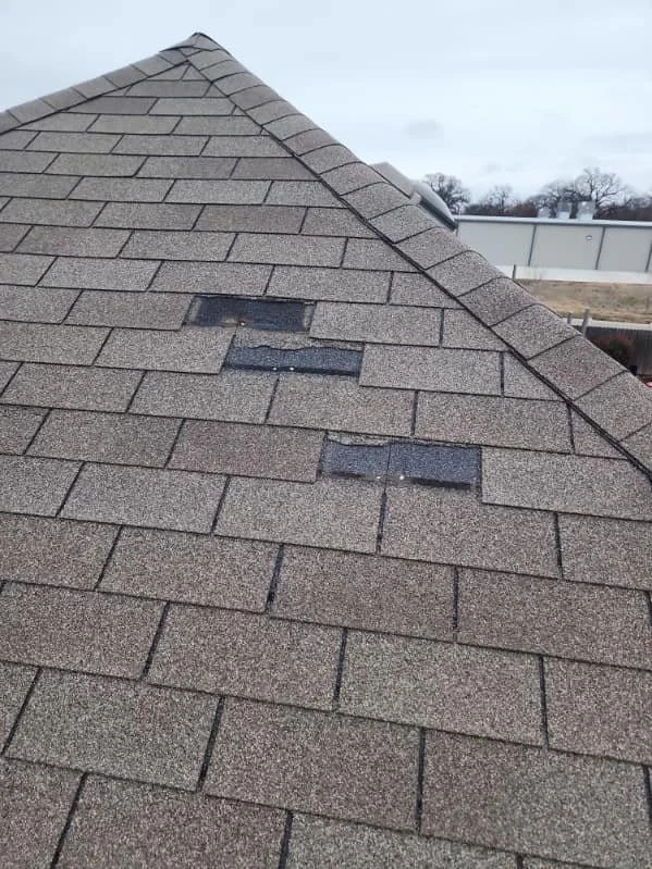 Close-up of a roof with asphalt shingles, showing two damaged shingles with black patches.