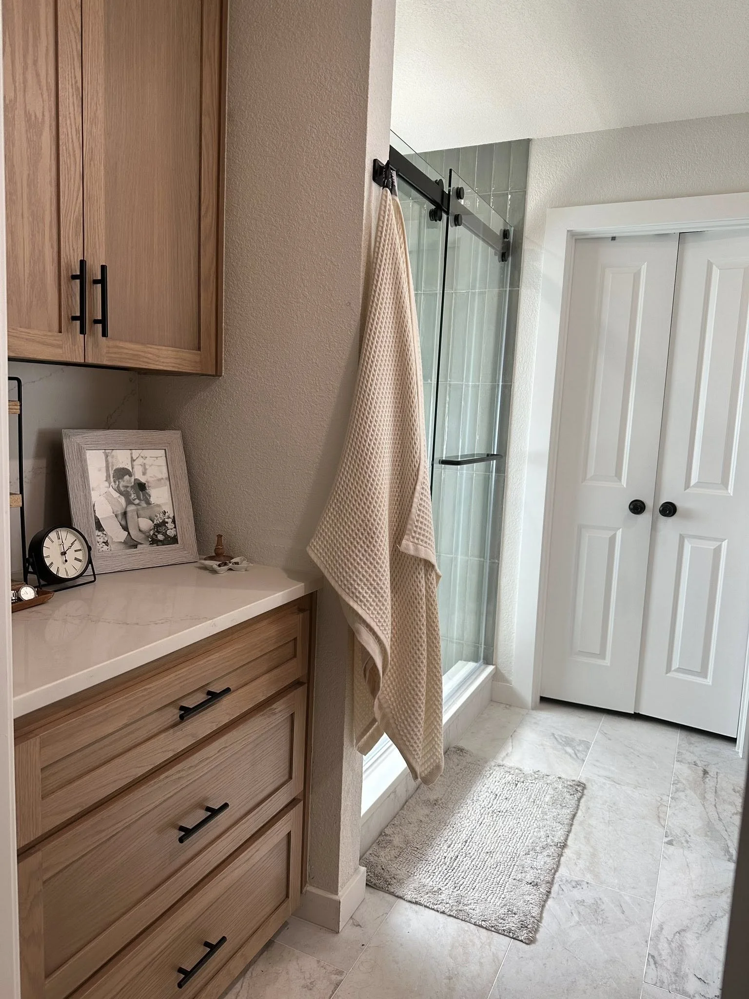 A bathroom with a corner shower with clear glass sliding door, beige towels hanging, wooden cabinets, and a gray rug on light-colored tile floor.