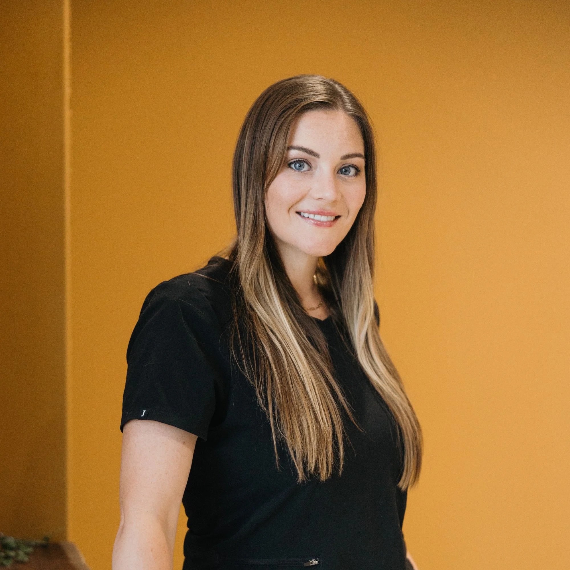 A woman with long wavy reddish-brown hair, wearing a black shirt and small hoop earrings, smiling in front of a plain light blue background.