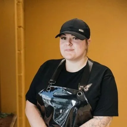 A woman with shoulder-length brown hair, wearing a black T-shirt with a skeleton and the word "NEVER", standing against a light blue wall.