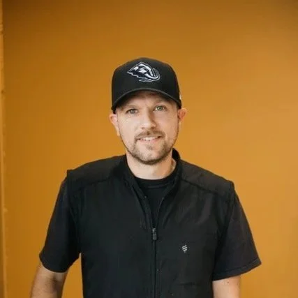 A man sitting with hands folded, wearing a black vest, wristwatch, and sitting on a turquoise chair. Behind him are framed wall art and a tapestry.