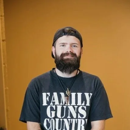 A man with a beard and tattoos sitting in a blue chair in front of a beige wall with framed art, including a Utah hockey banner, a cartoon pig, and a colorful dragon-themed tapestry.