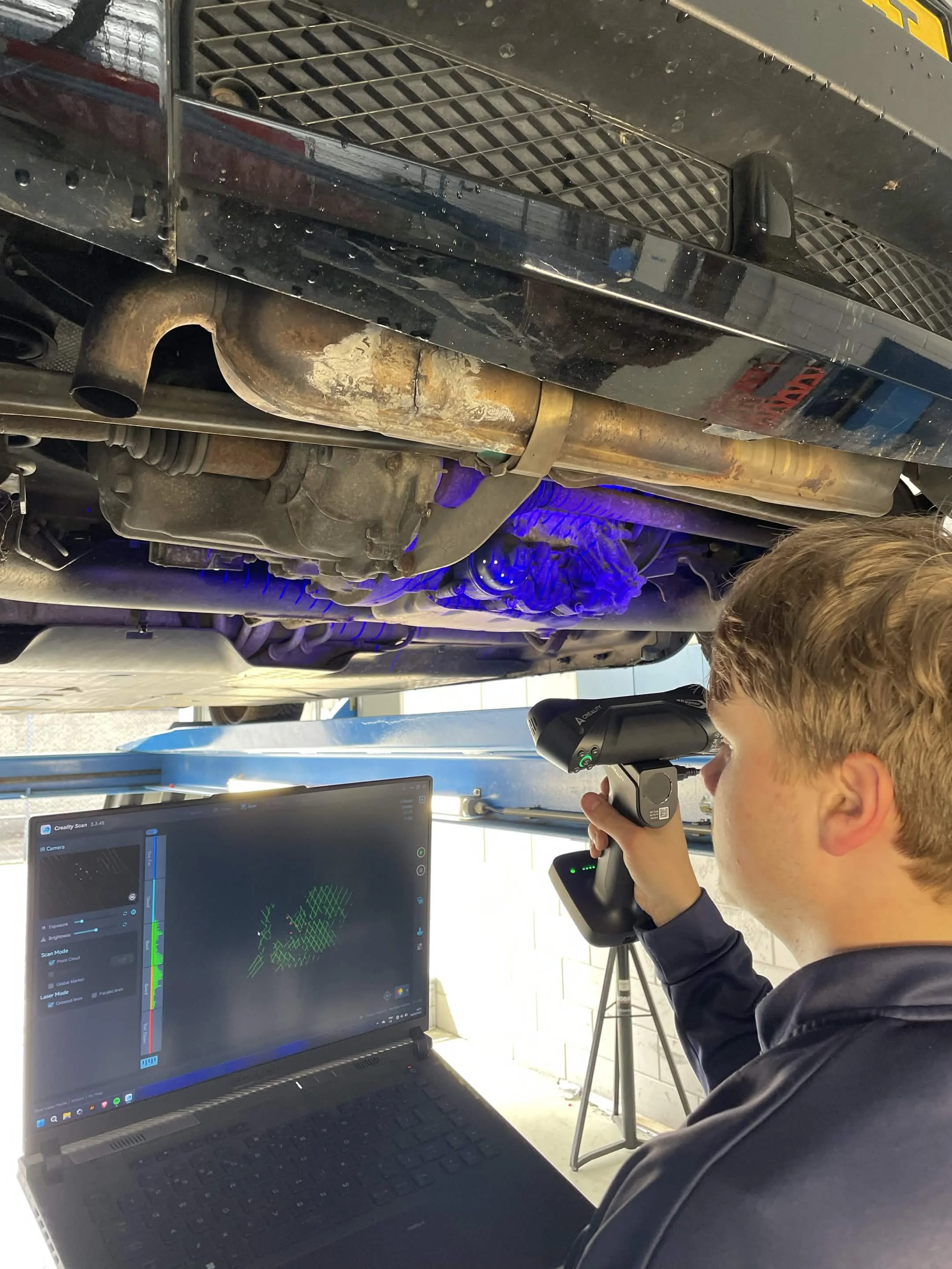 Een jonge man gebruikt een apparaat om de onderkant van een auto te scannen terwijl hij naar een computerscherm kijkt dat verschillende grafieken en gegevens toont.