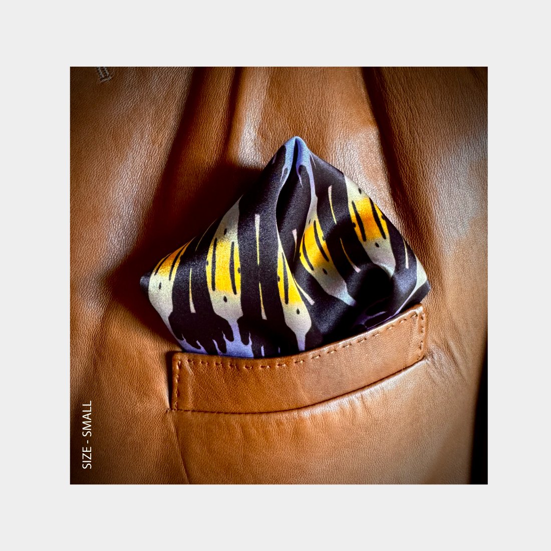 "Sunset Sentinels" is shown as a silk Pocket Square flowering with brilliant oranges, yellows and purples from the breast pocket of a brown leather jacket.