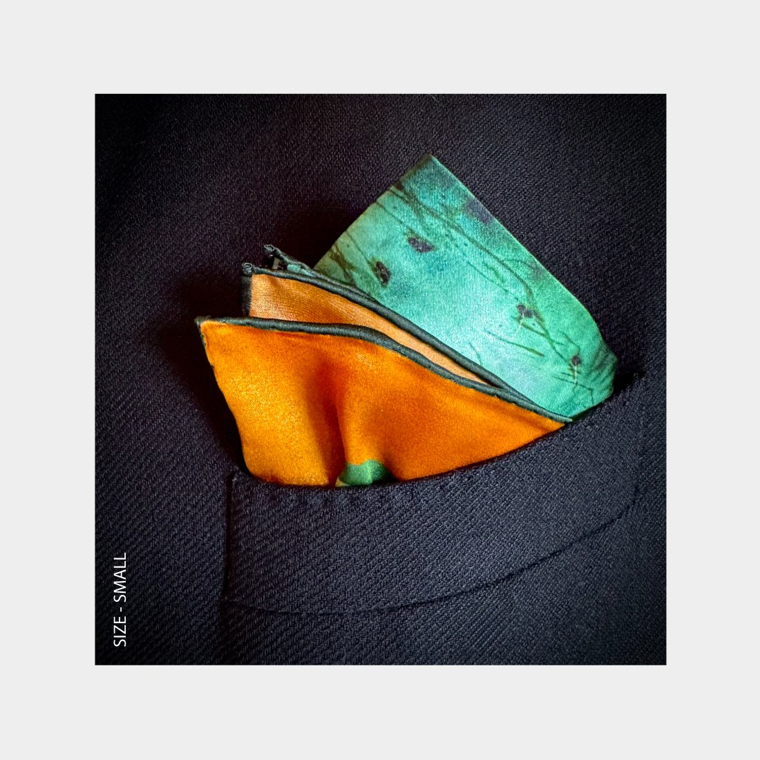 "Quenched" is shown is a Pocket Square folded as a pop of teal and orange pattern in the breast pocket of a black wool jacket.
