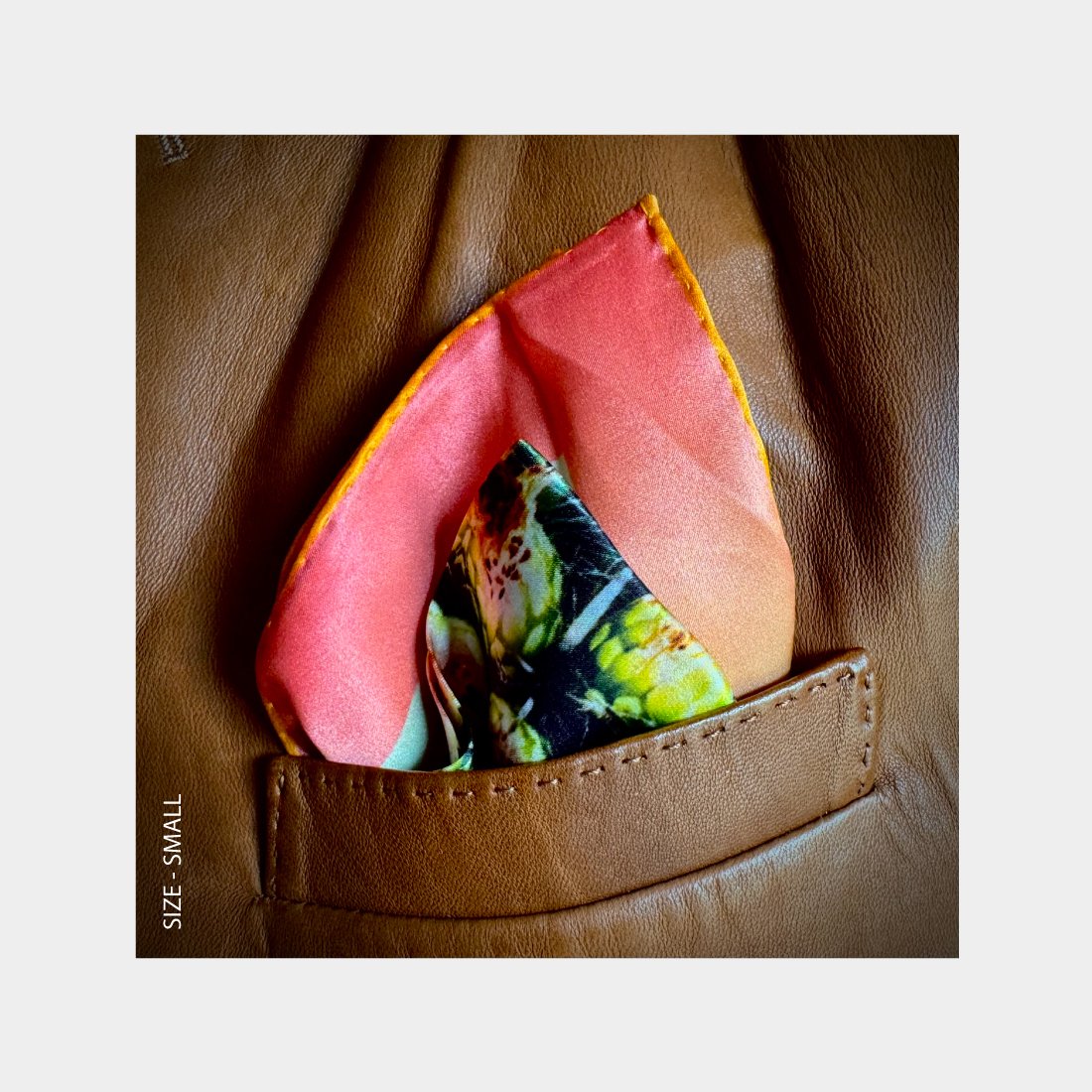 "Web of Life" is shown as a silk Pocket Square flowering with brilliant pinks, greens, and yellows from the breast pocket of a brown leather jacket.