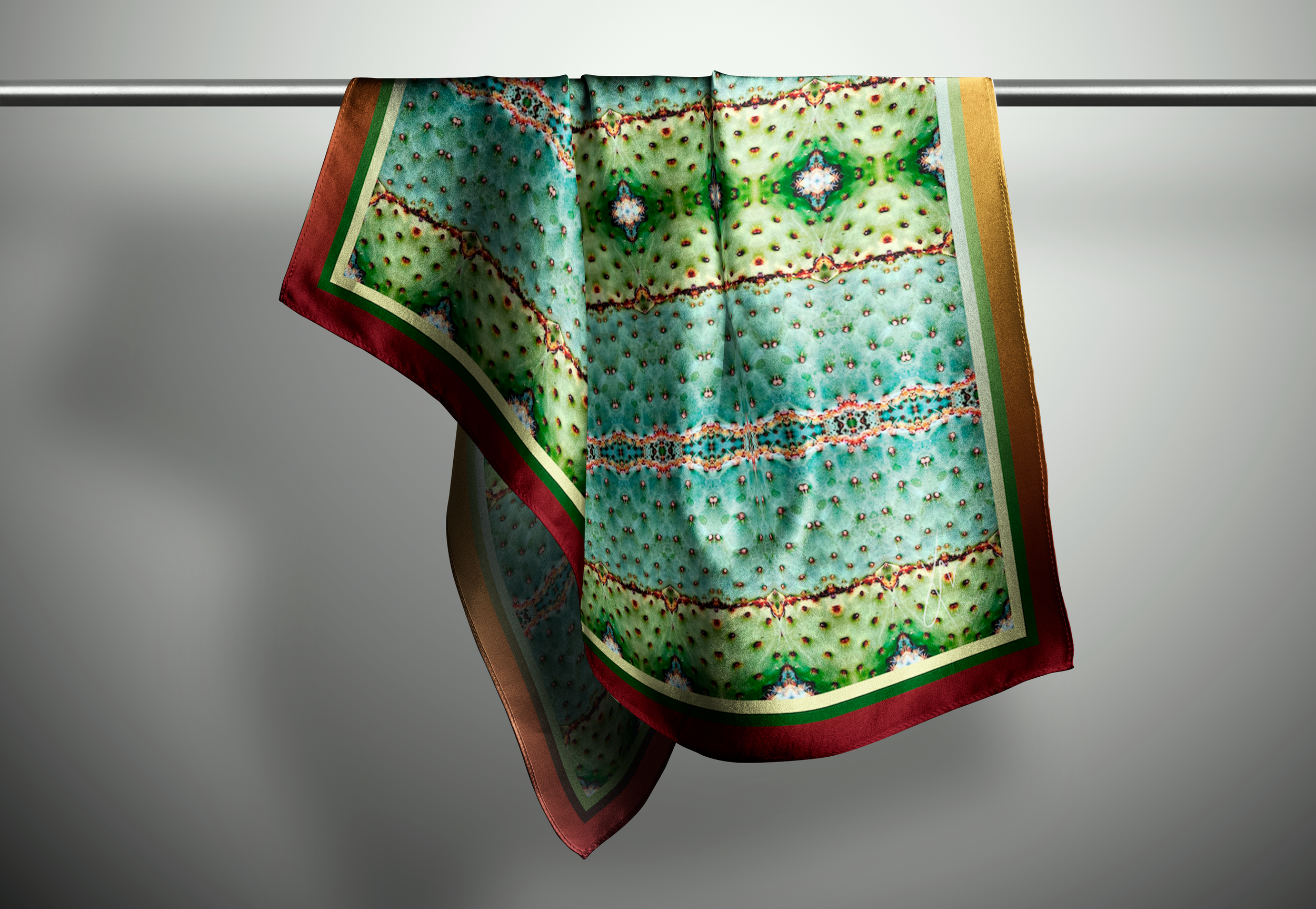 a picture of an intricate botanical patterned scarf in green, teal, and orange colours hanging on a rod within a light neutral background.