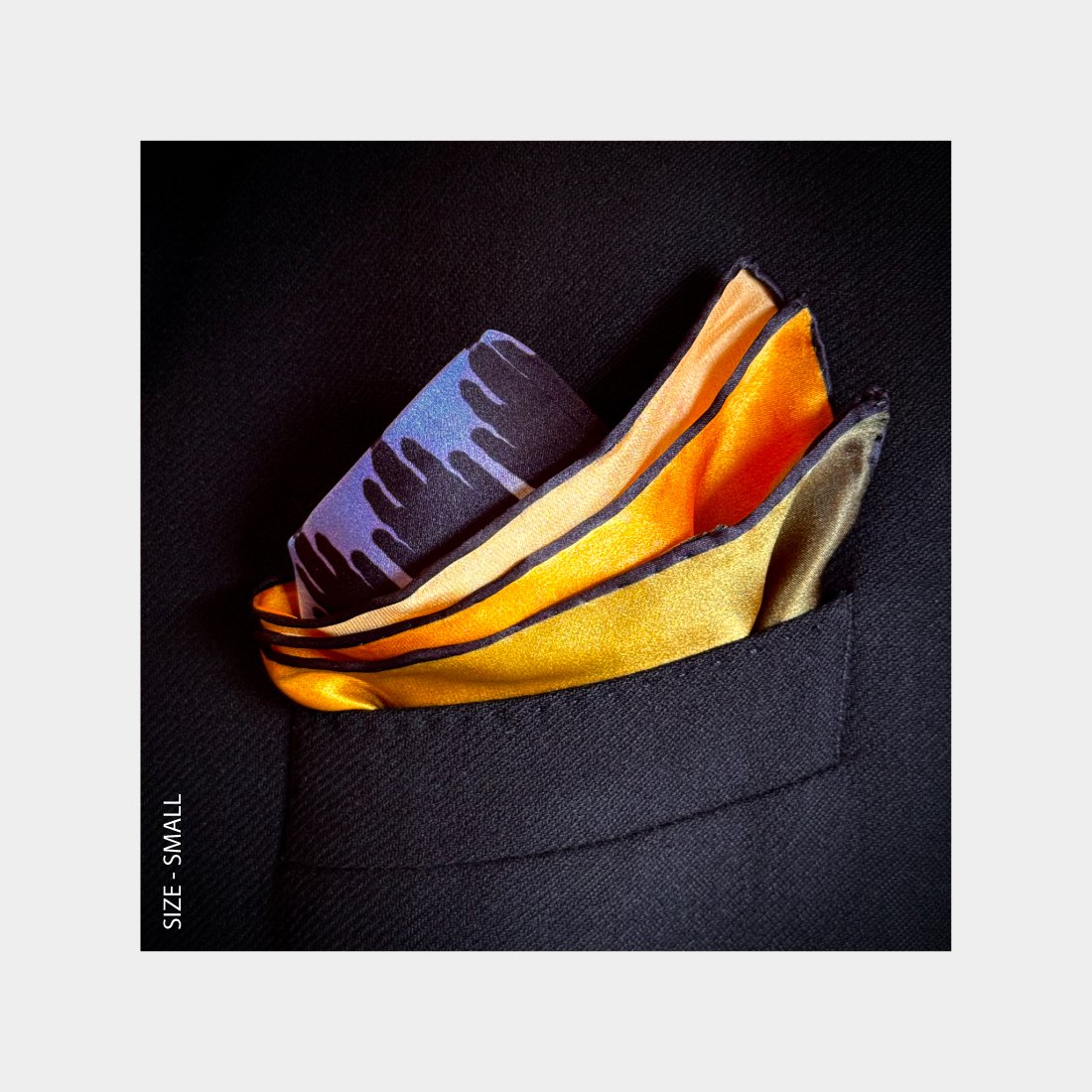 "Sunset Sentinels" is shown as a silk Pocket Square flowering with brilliant oranges and purples from the breast pocket of a black wool jacket.