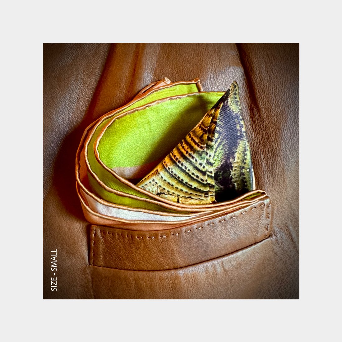 "Their Roots Grow Deep" is shown as a silk Pocket Square bursting with brillant green and earthy tones from the breast pocket of a brown leatherjacket.