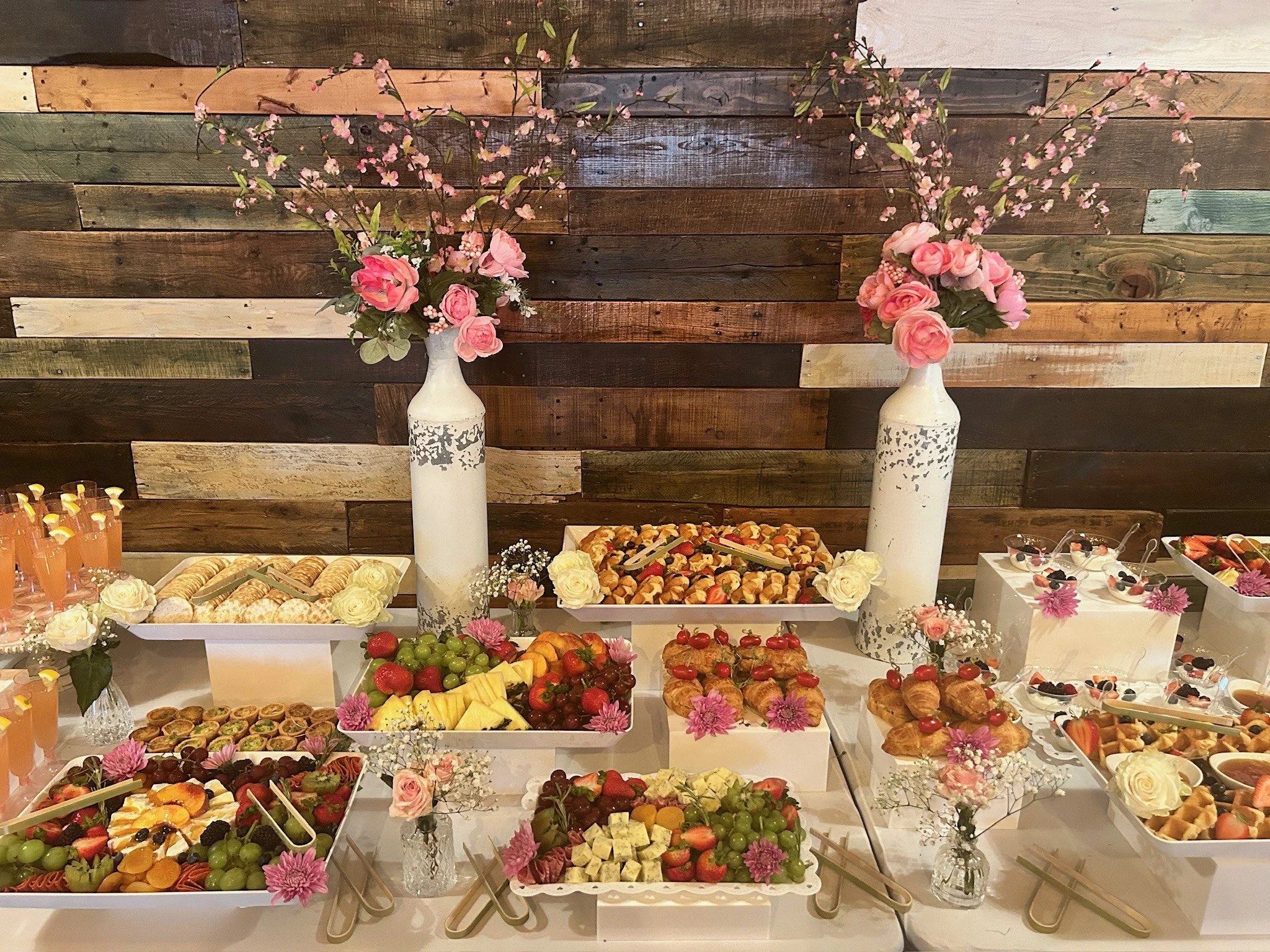 A dessert and fruit table decorated with pink and white flowers, featuring assorted pastries, fruit platters, and desserts in white trays and bowls against a wooden wall background.