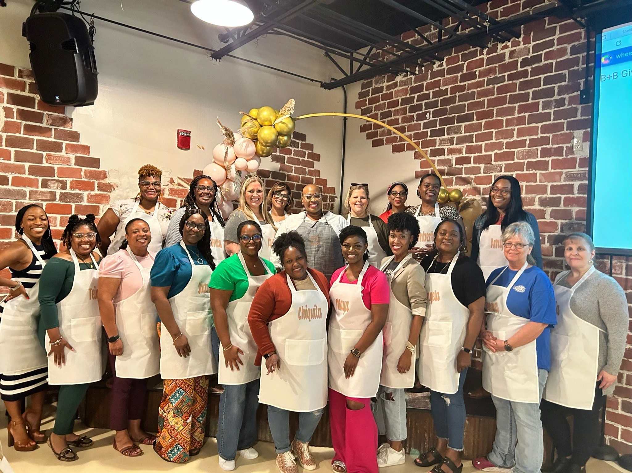 Group of women standing together in a room with a brick wall background, wearing aprons, celebrating an event, with balloon decorations above them.