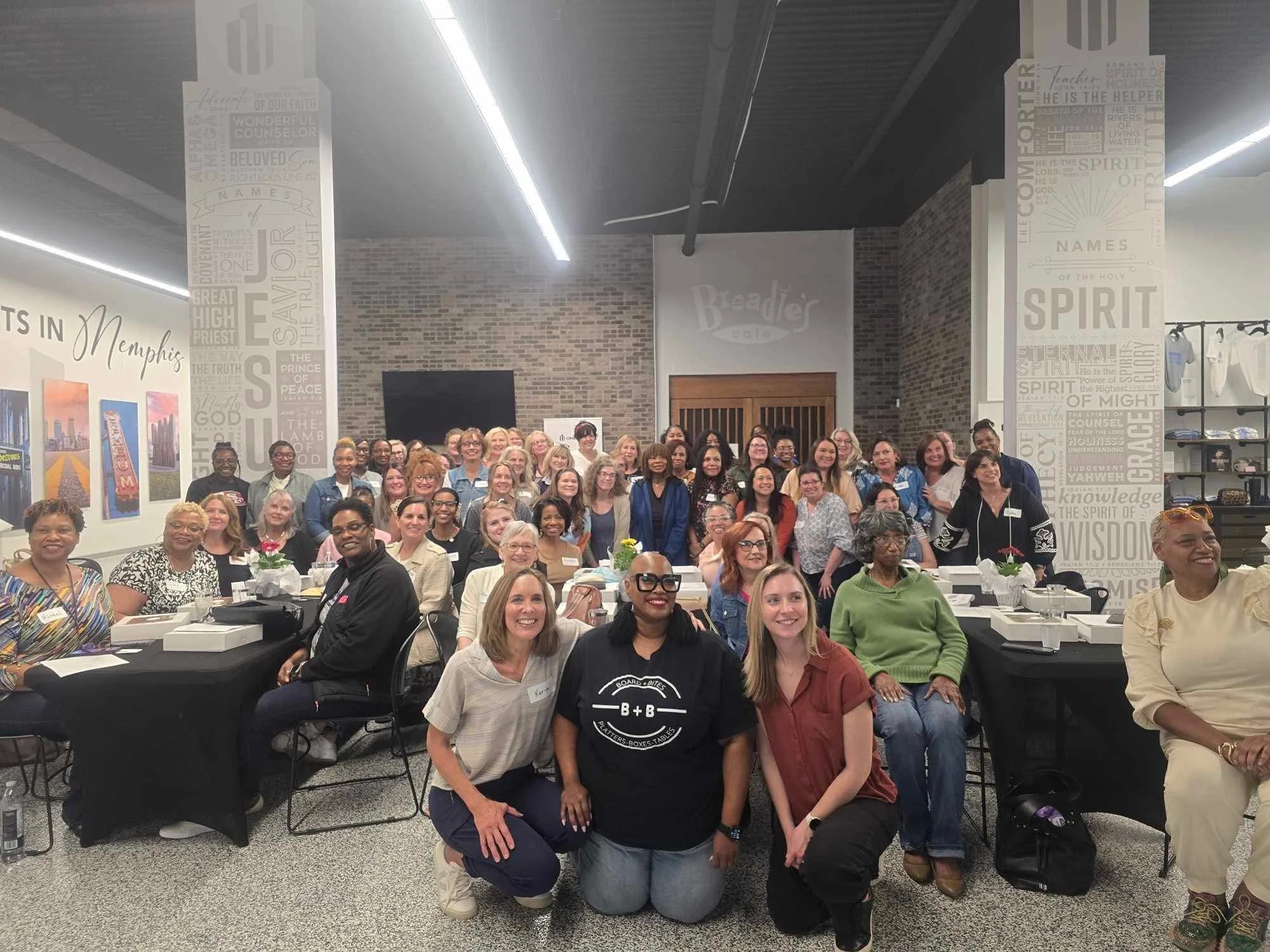 A large group of women and a few men gathered in a conference room, smiling for a group photo. The room has brick walls, black tables, and decorative text on the walls.