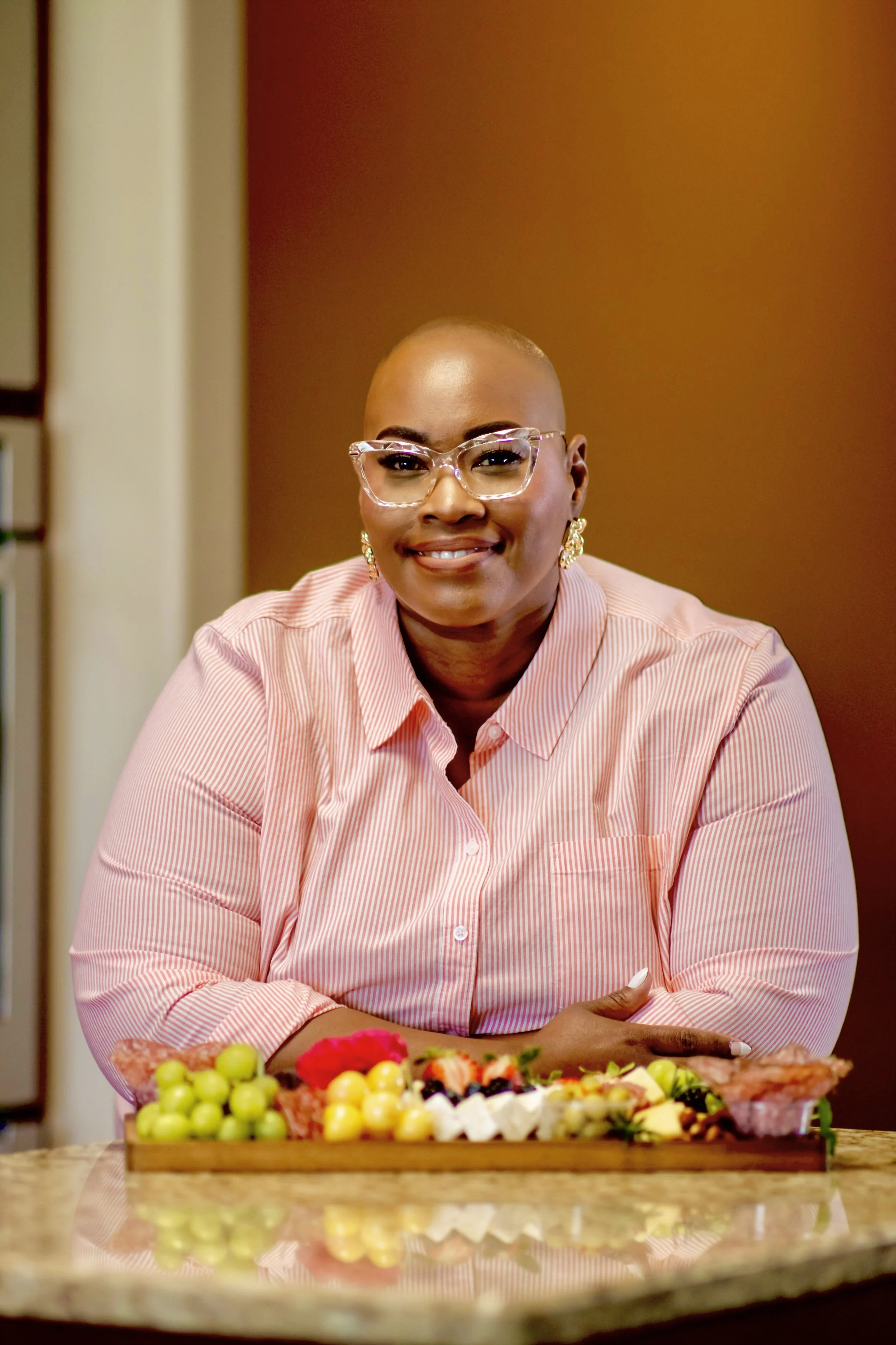 A woman with glasses and earrings wearing a pink pinstripe shirt, sitting at a kitchen counter with a tray of fruits and snacks in front of her, smiling at the camera.